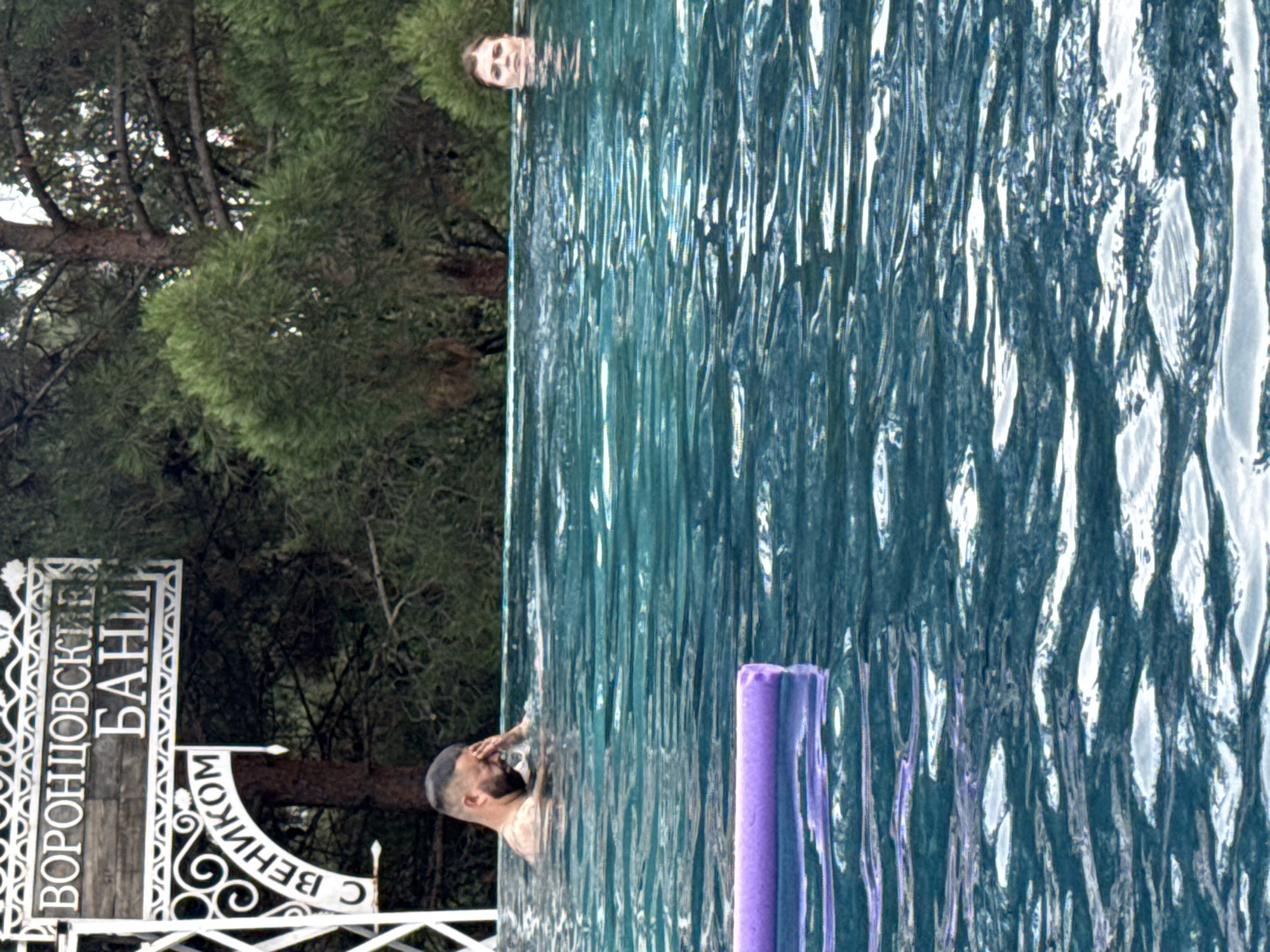 This image shows an outdoor swimming pool with two people in it, a man and a woman. The man is on the left side of the frame, with his head and shoulders visible above the water. He has a beard and is touching his forehead with his hand. The woman is on the right side of the frame, with her head and shoulders also visible. She has light-colored hair. A purple pool noodle is floating on the water in the foreground. In the background, there is a sign with Cyrillic lettering that appears to read "ВОРОНЦОВСКИЕ БАНИ" and "С ВЕНИКОМ", which translates to "Vorontsovsky Baths" and "With a Broom". The setting appears to be a forested area.