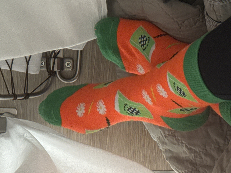 An indoor scene, depicting a pair of human feet, is observed in Gryazi, Russia. The feet are clad in matching bright orange socks with distinct green cuffs, heels, and toes. The socks feature a repeating pattern of white daisy-like flowers with yellow centers, thin black lines with yellow segments, and green geometric flag-like shapes containing a black hashtag symbol. Dark-colored trousers or leggings are visible just above the sock cuffs. The feet rest on a quilted grey fabric surface, likely a blanket or upholstery. In the background, a light brown surface exhibiting a wood grain texture is visible, along with sections of white fabric. A polished metallic fixture, possibly a bed frame component or a bracket, is also present in the upper right, secured by visible screws. The scene is evenly lit.