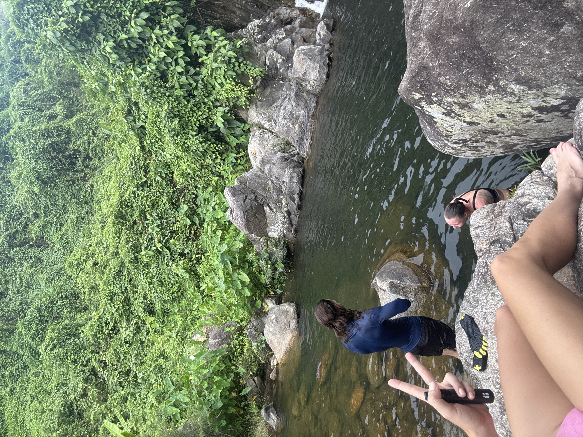 A scene in Đà Nẵng, Vietnam. Lush green vegetation covers a rocky terrain leading to a natural pool. Two people are partially submerged in the water. One is facing into the pool, wearing a blue long-sleeve top and dark shorts, while the other is further out, wearing a dark swimsuit. The right side of the image features legs and feet of an individual sitting on a rock, holding a small black object. Another person's hand is visible, displaying a peace sign.