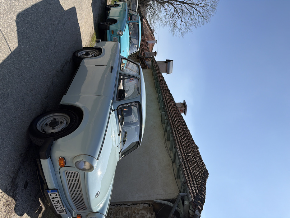 The image displays a clear daytime scene in Bukovets, Bulgaria, featuring multiple vehicles parked on an asphalt road adjacent to residential structures. In the foreground, positioned to the right, is a light blue, two-door Trabant 601 sedan. Its front license plate, "CA 2922 TM," is visible. Directly behind this vehicle, further down the road and to the left, a second, turquoise-colored Trabant 601 sedan is parked. A third, partially obscured white vehicle is visible in the distant background.
To the right of the foreground car, a building with light-colored, rendered walls and a steeply pitched roof covered in dark terracotta tiles is present. A brick chimney and a smaller metal vent are visible on the roof. The lower right section of the building's facade reveals exposed stone masonry and a prominent vertical crack. On the far left, a large, leafless deciduous tree with extensive branching suggests a late winter or early spring season. The sky is clear and blue, with no clouds. Strong sunlight casts distinct shadows of the vehicles and other objects onto the road surface. No individuals are present in the depicted scene, and no actions or interactions are occurring.