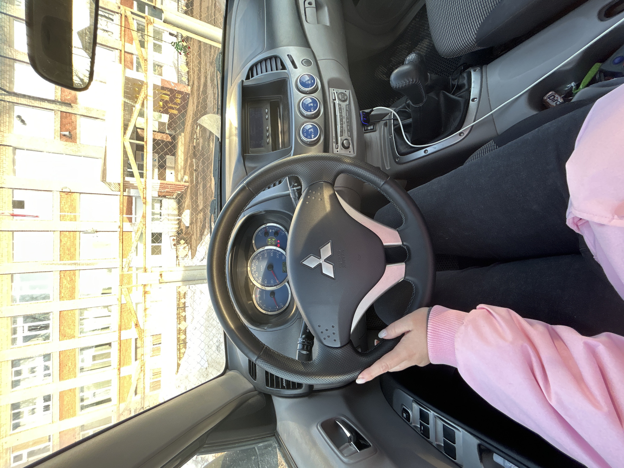 The image shows the interior of a car, specifically the driver's seat. The steering wheel, bearing a Mitsubishi logo, is prominently featured. A person wearing a pink jacket is visible, with their hand resting on the steering wheel. The car's dashboard, including the instrument panel and infotainment system, is also visible. The view through the windshield reveals a building with scaffolding outside, possibly indicating construction or maintenance work. The location appears to be Фоки, Russia.