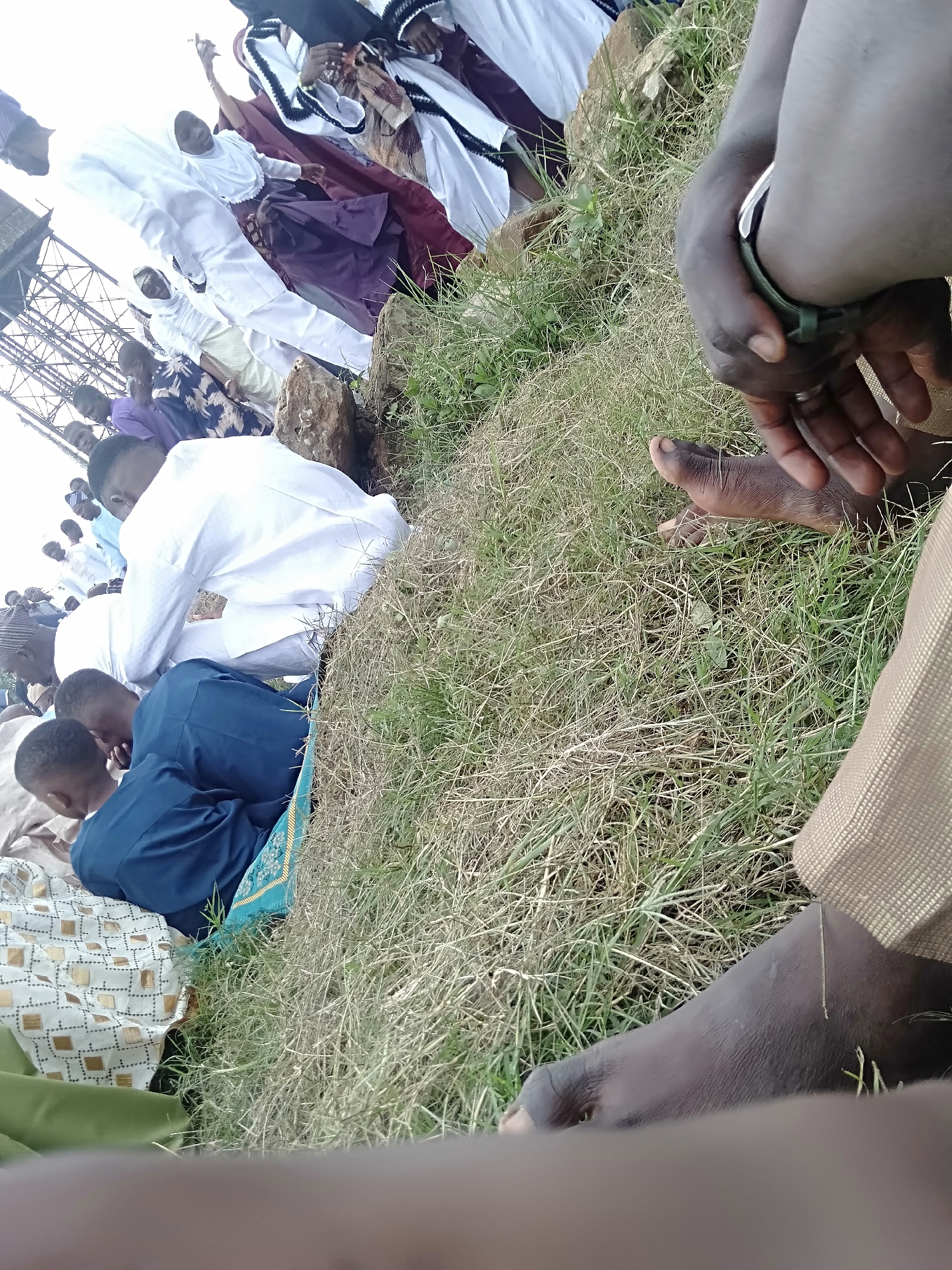 A low-angle shot shows a group of people gathered on a grassy area in Unknown, Unknown. Most of the people in the image are wearing white clothing, while others are wearing darker clothing, including blue and purple. One man kneels on what appears to be a prayer rug or patterned cloth. The foreground is obscured by a partially visible person's arm and leg. In the background is a tall metal structure, possibly a tower. The people in the background appear to be standing. The overall scene is somewhat unorganized.
