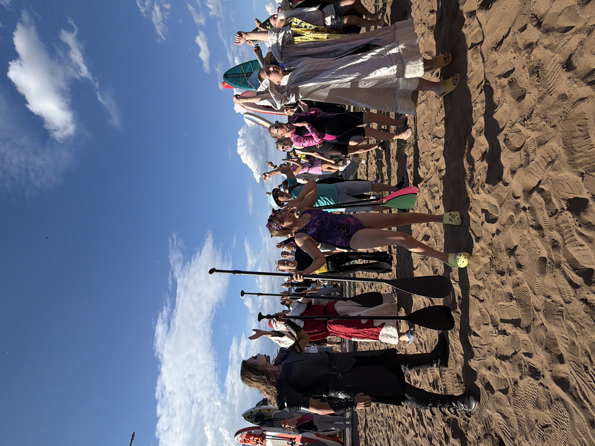 A group of people are gathered on a sandy beach, standing in a line, many holding paddles and some with what appear to be surfboards or paddleboards. The sky is a bright blue with scattered white clouds. The sun is shining, casting shadows on the sand, suggesting it is daytime. The people are dressed in a variety of outfits, some in swimsuits and athletic wear, while others are in costumes, including what appears to be a person dressed as a zombie at the front of the line. Some individuals are wearing sandals or flip-flops. The scene appears to be festive and possibly part of an organized event or activity. There is no visible text in the image.