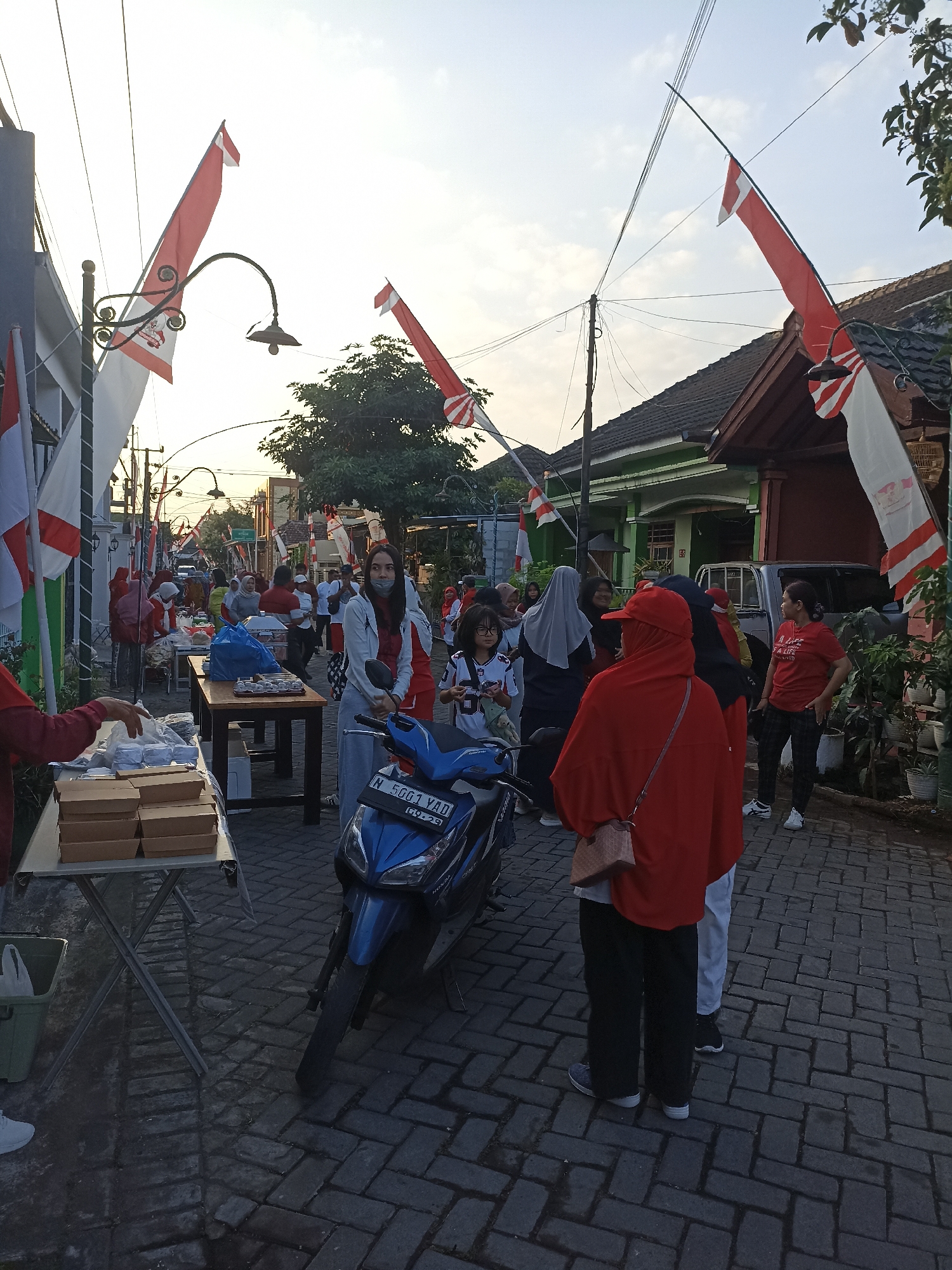The image depicts a street scene in Sumberejo, Indonesia, likely during a festive occasion given the abundance of red and white Indonesian flags displayed on poles and strung across the street. The street is paved with interlocking brick pavers. Several people are visible, some standing in small groups, others interacting with vendors.
In the foreground left, a table is laden with stacked cardboard boxes, suggesting a food stall or distribution point. A person in a red shirt is partially visible on the far left, with their hand extended towards the table.
To the right of the table, a blue motorcycle is parked, its license plate reading "N 5001 YAD". A young girl with glasses and a white and red top stands beside the motorcycle.
Further into the scene, people are gathered around tables where food or goods appear to be laid out. Some individuals are wearing red shirts, matching the national colors. A woman in a grey hijab and a white and red top is engaged in conversation with another person. Behind them, a woman in a red headscarf and red outer garment is facing away from the camera, with a light brown patterned bag slung over her shoulder.
A silver van or small truck is parked in the background, near a building with a green exterior and a dark roof. Houses with red tile roofs line the street. The sky is bright, indicating daytime, possibly early morning or late afternoon, with a soft glow. The overall atmosphere suggests a community gathering or event.