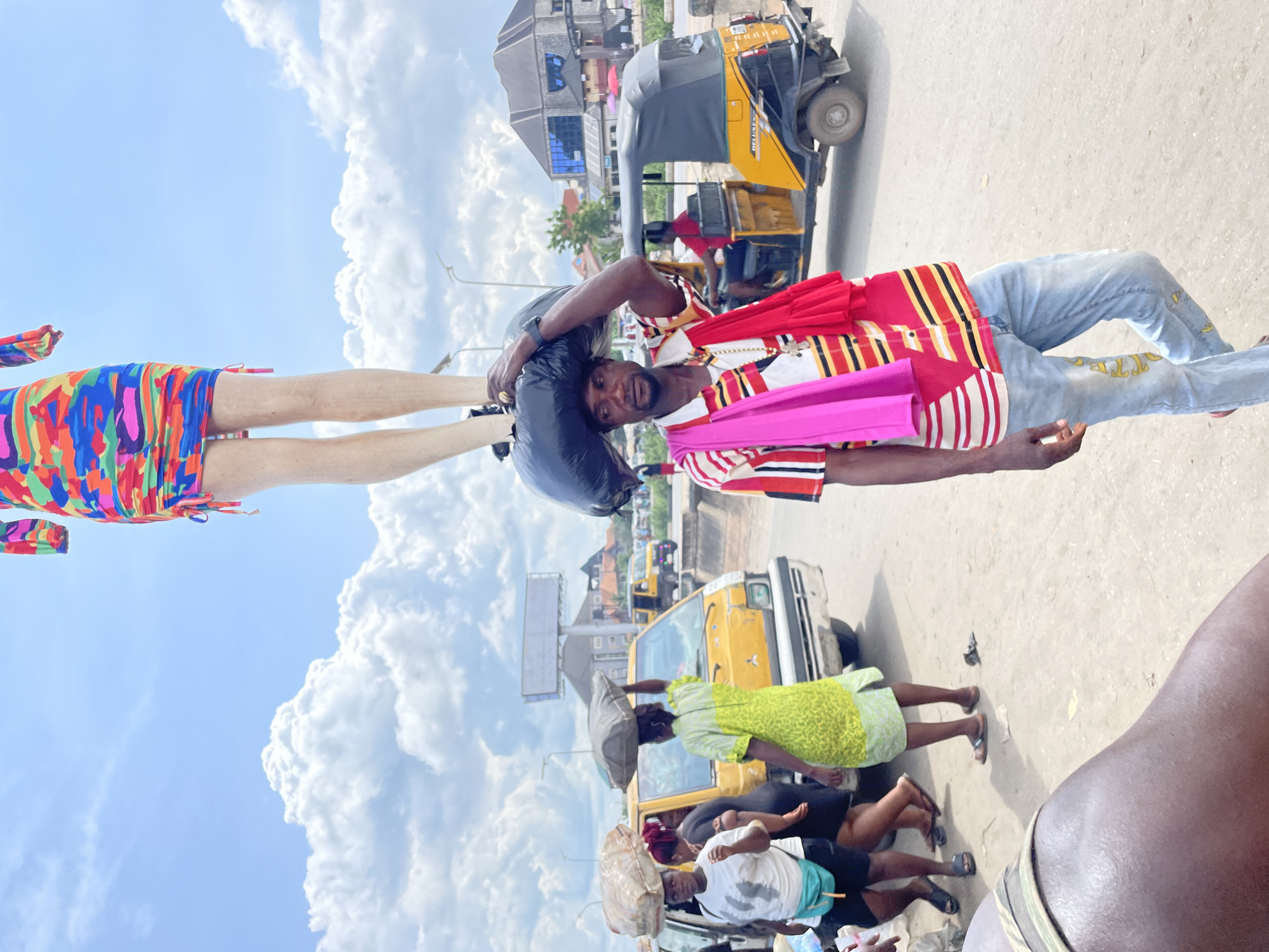 The image depicts a bustling outdoor street scene, likely in a West African city, under clear, bright daylight with a blue sky and scattered white clouds. The photograph is rotated 90 degrees counter-clockwise.
The central subject is a dark-skinned man, captured from the waist up, looking directly at the viewer with a neutral expression. He wears a striking striped shirt in red, white, black, and yellow/gold, light blue jeans adorned with yellow patterns, and a dark watch. A black bag rests on his left shoulder/head, and he has red and bright pink fabrics draped over his shoulders.
To the left (when upright), against the sky, are the lower legs and torso of another person wearing a brightly colored, abstract-patterned dress in hues of blue, green, red, yellow, pink, and orange.
In the mid-ground, a yellow and black tricycle (Keke Napep or tuk-tuk) with "BELLVIEW" visible on its side is prominent. Behind it, a yellow commercial van can be seen. Several other individuals are present in the background: one woman in a light green patterned dress carries a package on her head, and another person in a white t-shirt also balances an item on their head. Buildings line the street, and a light pole stands against the sky. The ground appears to be a light-colored, dusty road surface. The scene portrays typical daily activity in a busy urban environment.