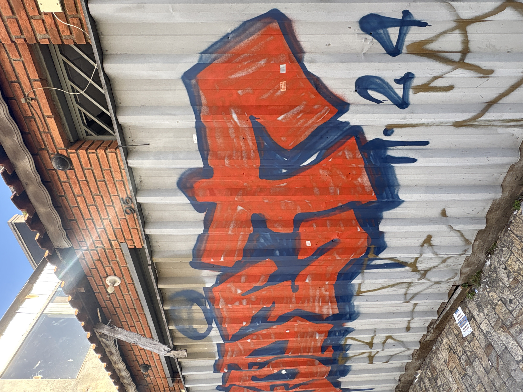 An outdoor, eye-level shot captures a corrugated metal wall covered in graffiti, possibly in a back alley or industrial area within São Paulo, Brazil. The wall is painted a metallic gray and features large, stylized graffiti tags in bright orange with blue outlines and fills. Some of the graffiti includes drips from the paint. To the right of the graffiti, the numbers "124" are spray-painted in blue. Fainter, older graffiti in a brown spray paint is visible underneath the larger orange and blue tags. Above the corrugated wall, a section of red brick wall with a tiled roof is visible. A window with diagonal bars is set into the brick wall. The ground in the foreground is paved with cobblestones and scattered with debris, including a crumpled piece of paper. The lighting suggests it is daytime, with some sunlight casting shadows and creating lens flare in the upper left portion of the image.