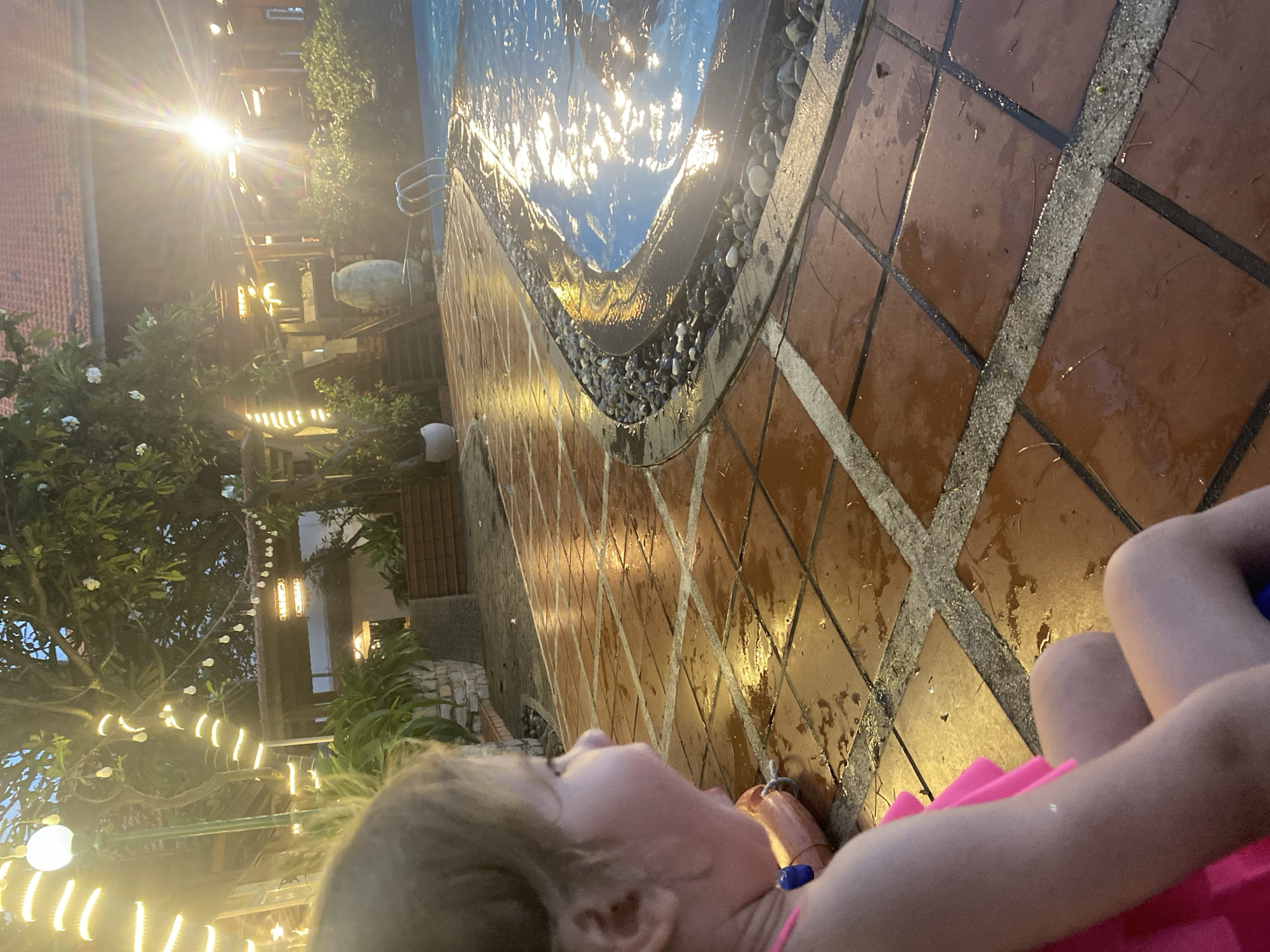 This image, tilted to the side, captures an illuminated outdoor scene at dusk or night in Phu Thuy Ward, Vietnam, centering on a child near a swimming pool. In the immediate foreground, a young child, likely a toddler, is partially visible from the chest up, wearing a bright pink garment. The child's head is turned upwards and slightly to the left, seemingly observing the lit surroundings.
The child is situated on a reddish-brown tiled patio, which appears wet, with light-colored grout lines forming a grid pattern. These wet surfaces reflect the artificial lights, creating shimmering patches. The patio curves around a swimming pool, whose dark blue water is intensely reflective, mirroring numerous bright lights and appearing to glitter. The edge of the pool is adorned with a border of small, light-toned pebbles.
The background is dominated by artificial light sources. A powerful, starburst-like light emits a strong glare from the upper left. Various string lights with round bulbs are draped across trees and other foliage, casting a warm glow. Other illuminated structures, possibly parts of buildings or an outdoor seating area, are visible through the dense greenery. The overall atmosphere suggests a serene and inviting evening at a resort or hotel. There is no visible text in the image.