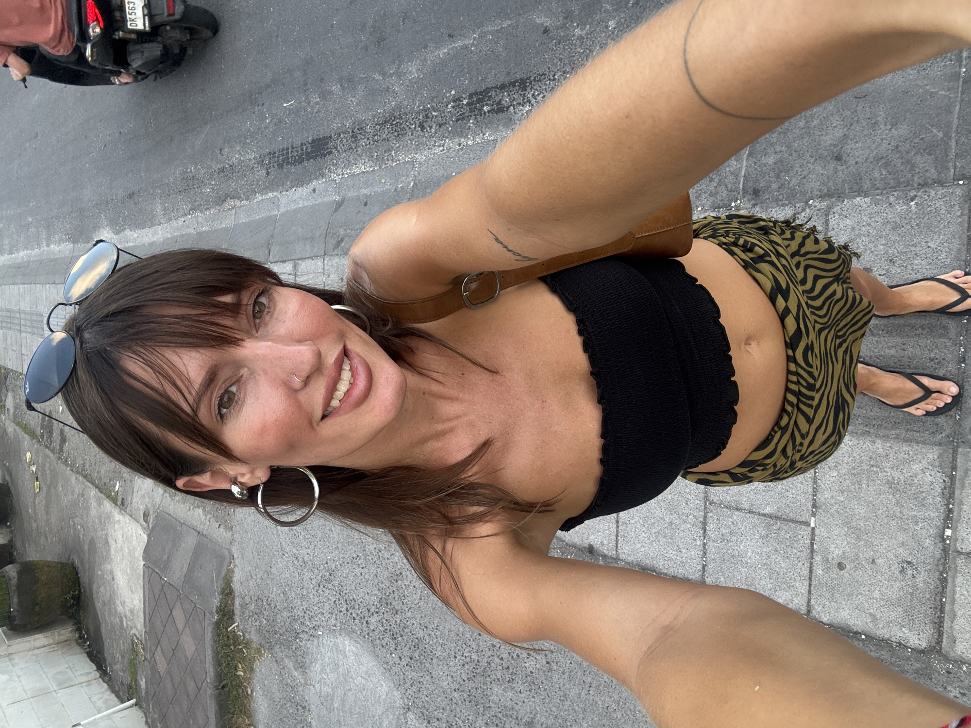 A woman is taking a selfie outdoors. She is wearing a black top, a patterned skirt, and flip-flops. Sunglasses are perched on her head, and she wears hoop earrings. A brown shoulder bag is slung over her shoulder. A motorcycle is visible in the background, parked on a grey asphalt road. The road appears to be next to a sidewalk made of grey concrete blocks. Based on the license plate visible on the motorcycle, the location is North Kuta, Indonesia.