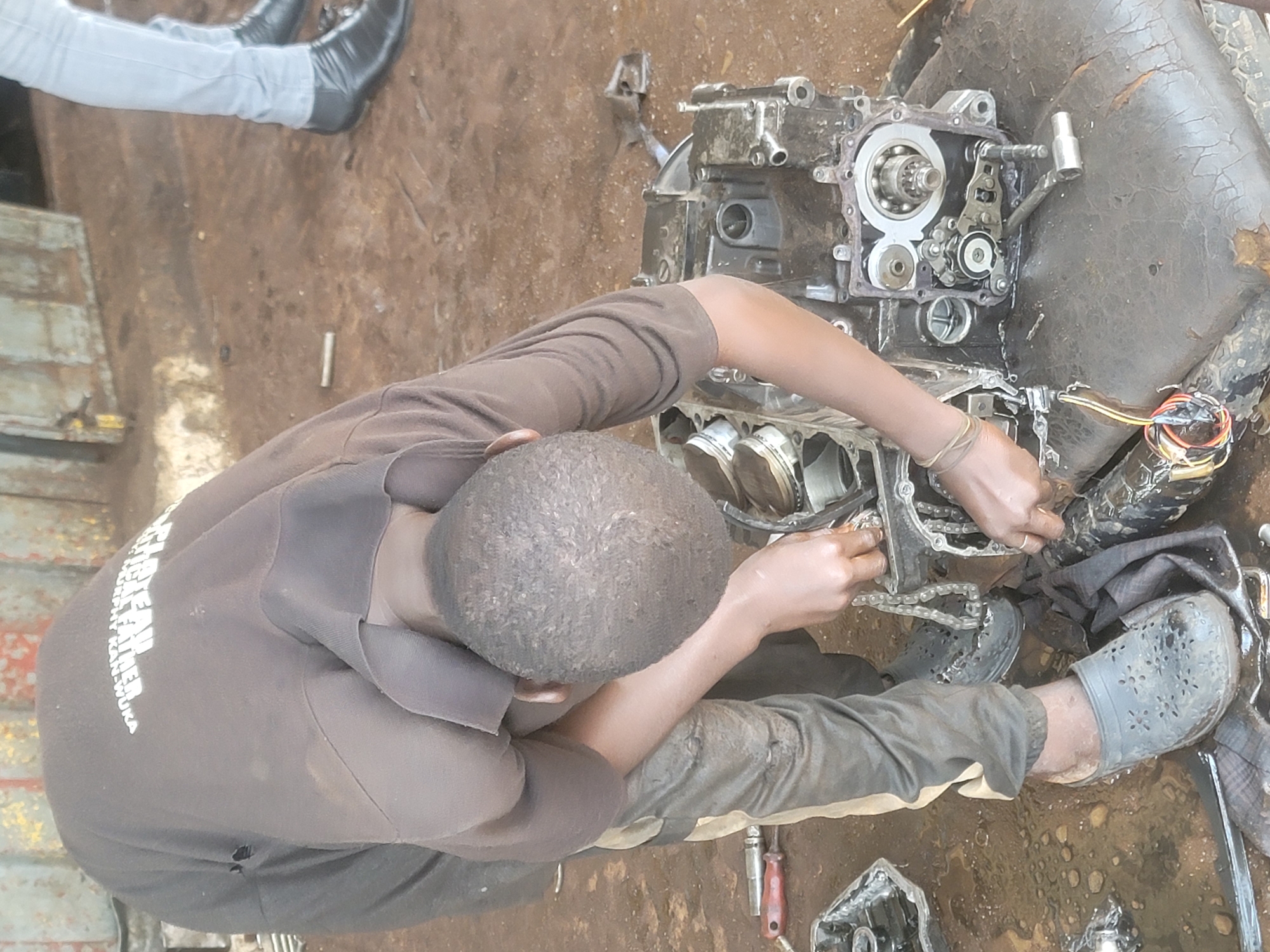 In Barakin Lerawi, Nigeria, a mechanic, hunched over, works on a dissembled motorcycle engine in a workshop. The ground is wet and littered with debris. The mechanic wears a dark brown t-shirt, dark pants with a tan stripe, and gray perforated clogs. A second person, whose legs are visible, wears light blue jeans and black leather shoes. The engine components are spread across a worn, dark leather surface. A bundle of colored wires is visible to the left of the engine. Tools, including a wrench with a red handle, are scattered on the ground. The workshop appears to be an open-air or semi-enclosed space, with metal structures in the background.