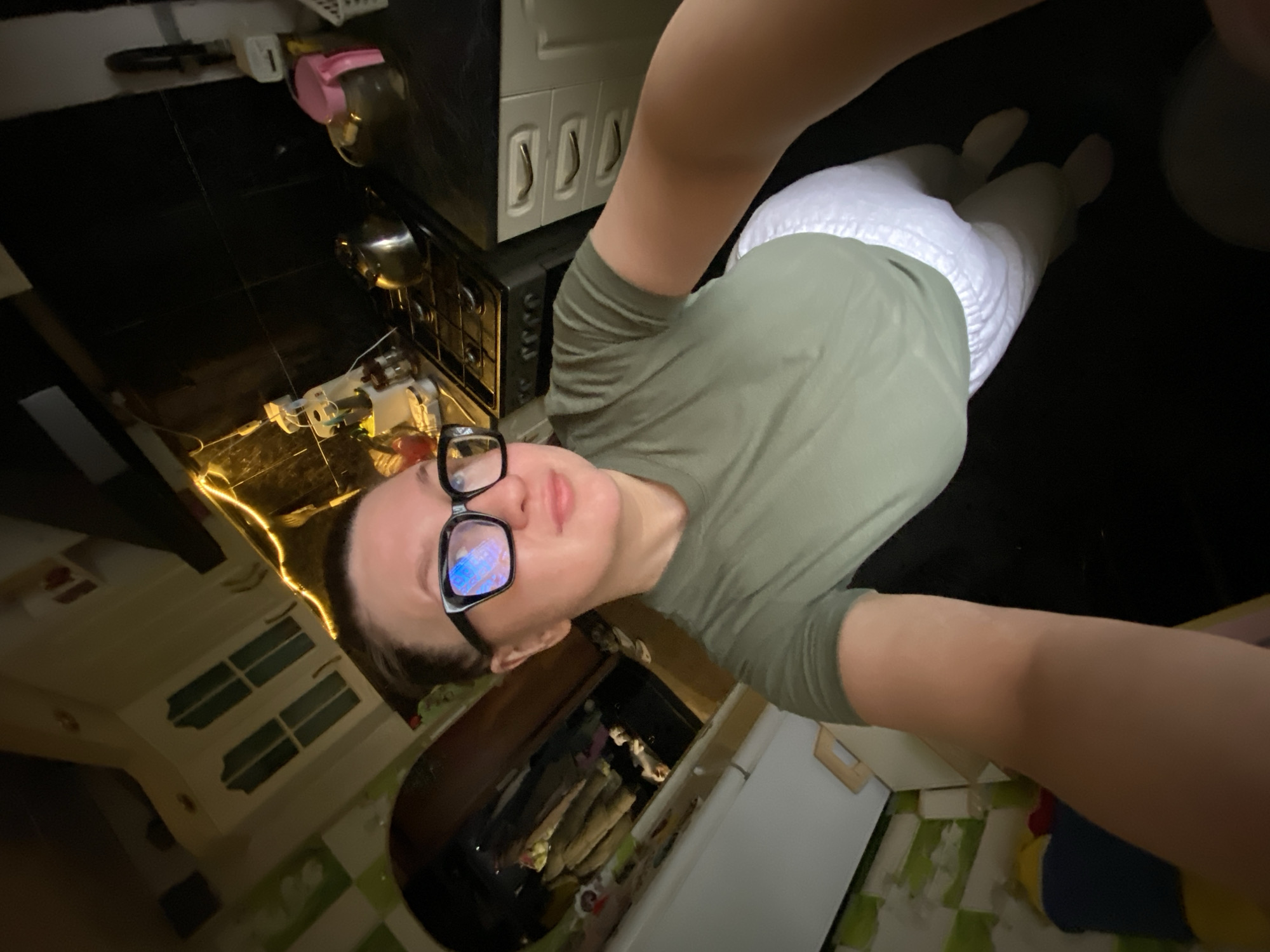 A selfie-style photograph depicts a person indoors, presumably in a kitchen. The person is wearing glasses and is positioned at an angle, with the camera held above. The person is wearing a green shirt and white pants.
The background includes kitchen elements such as countertops, cabinets, a stove, and kitchenware. The floor is tiled. The lighting in the kitchen appears to be a mix of natural and artificial light.
The location is unknown.