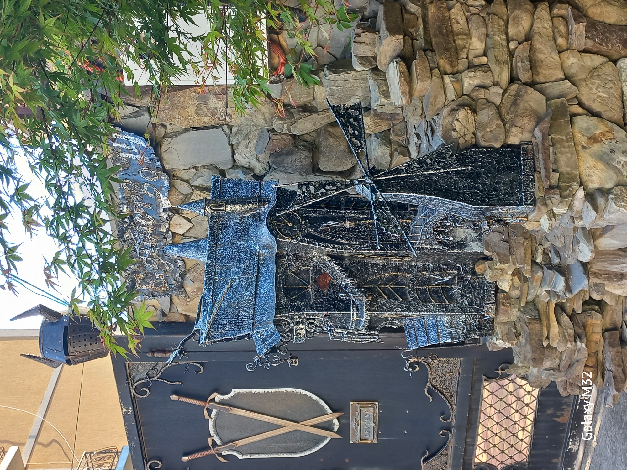 The image shows a gate in Sochi, Russia, adorned with various metallic decorations including a windmill, coat, shield with crossed swords, and letter box. The gate is positioned between a beige-colored building and a stone wall. Green foliage is visible above the gate.