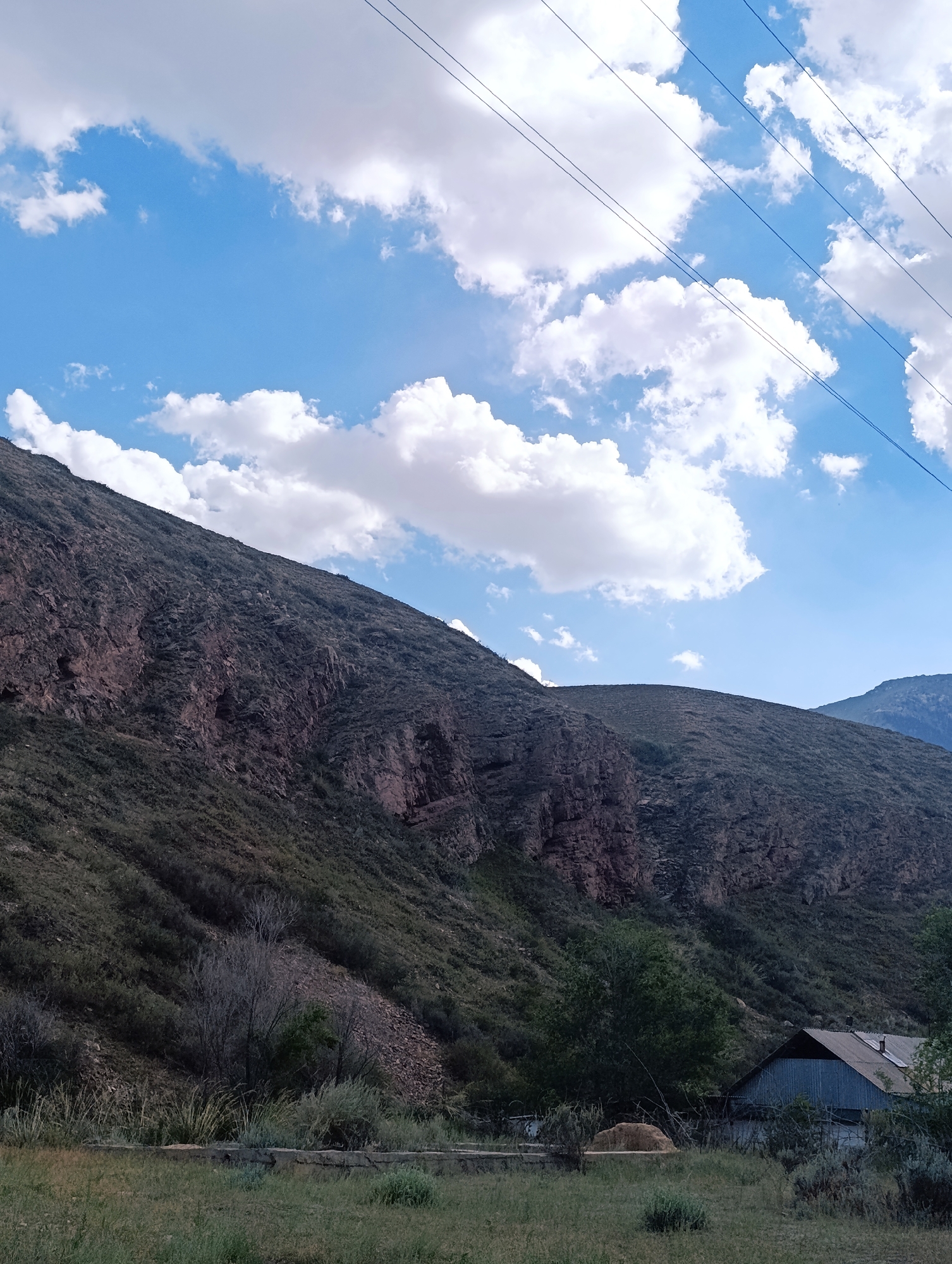 The image depicts a mountainous landscape with a visible building and power lines in a rural area. The foreground is dominated by dry grass and brush, with a concrete structure and a hay bale visible. A metal-roofed building with a chimney is situated to the right, partially obscured by trees and vegetation. The middle ground features a steep, rocky mountainside covered in sparse vegetation. The background consists of more distant, hazy mountains under a bright blue sky with scattered white clouds. Four parallel power lines stretch across the upper portion of the frame. The scene suggests a remote, possibly agricultural or residential, setting in a mountainous region. The location is identified as the city of Naryn, Kyrgyzstan.