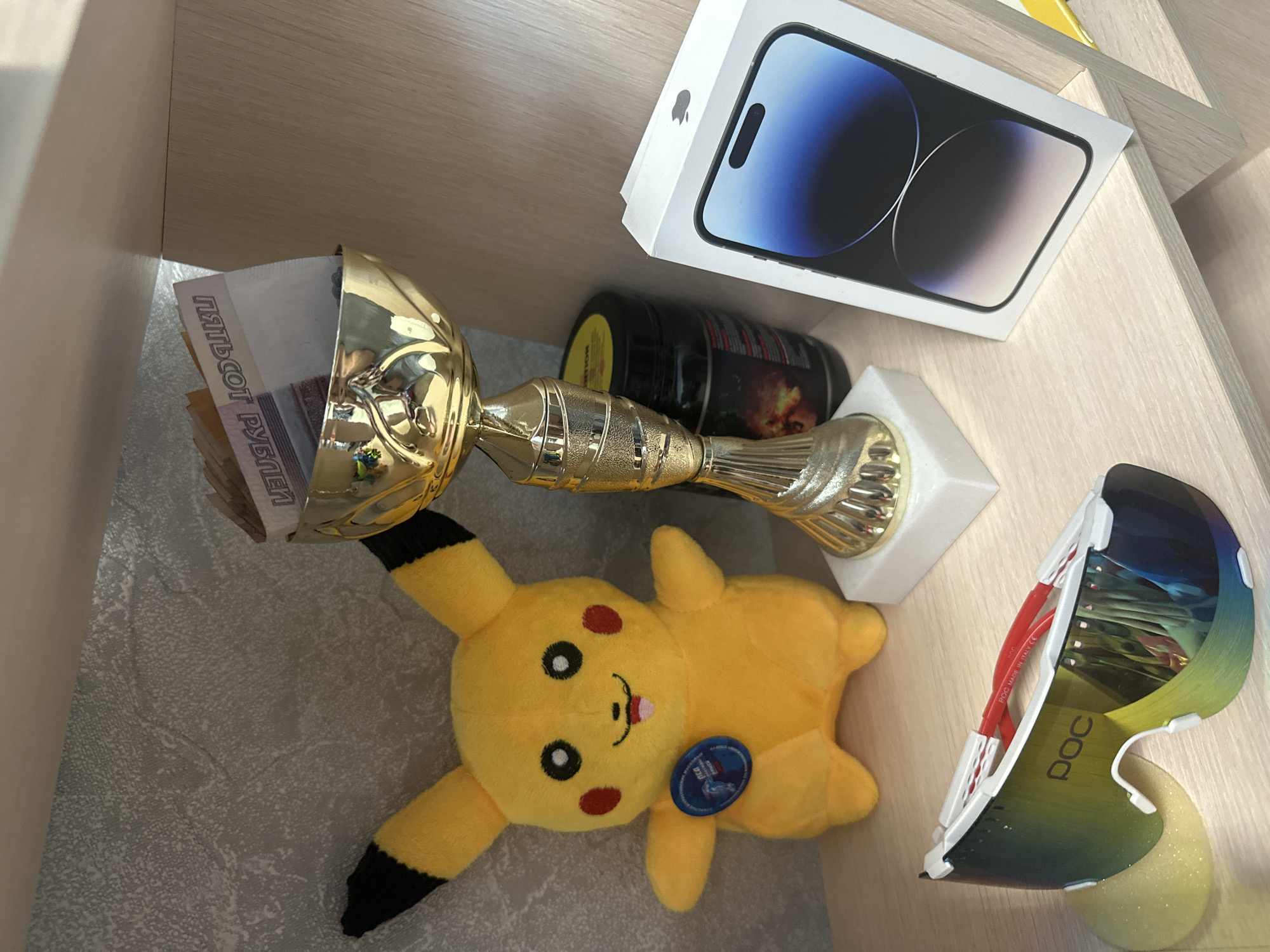 This image shows a collection of items on a wooden shelf. In the foreground, a plush yellow Pikachu toy is lying on its back. To its left, a golden trophy cup is tilted, with Russian banknotes, specifically 5000 ruble notes, visible inside. Behind the trophy and toy, a black cylindrical container with a red label is visible, and further back, a white box with an Apple logo and an image of a smartphone is placed. To the right of the Pikachu and trophy, a pair of white POC cycling glasses with mirrored lenses are resting on the shelf. The scene appears to be indoors, likely in a home or office, with soft lighting suggesting daytime. There is no explicit indication of the time of day or weather conditions. The items suggest a mix of personal interests and achievements, possibly related to sports, technology, or a gaming hobby. Some visible text includes "ПЯТЬСОТ РУБЛЕЙ" (five hundred rubles) on the banknotes, "POC" on the glasses, and "MADE IN ITALY CE" also on the glasses. The overall impression is one of personal belongings displayed together.