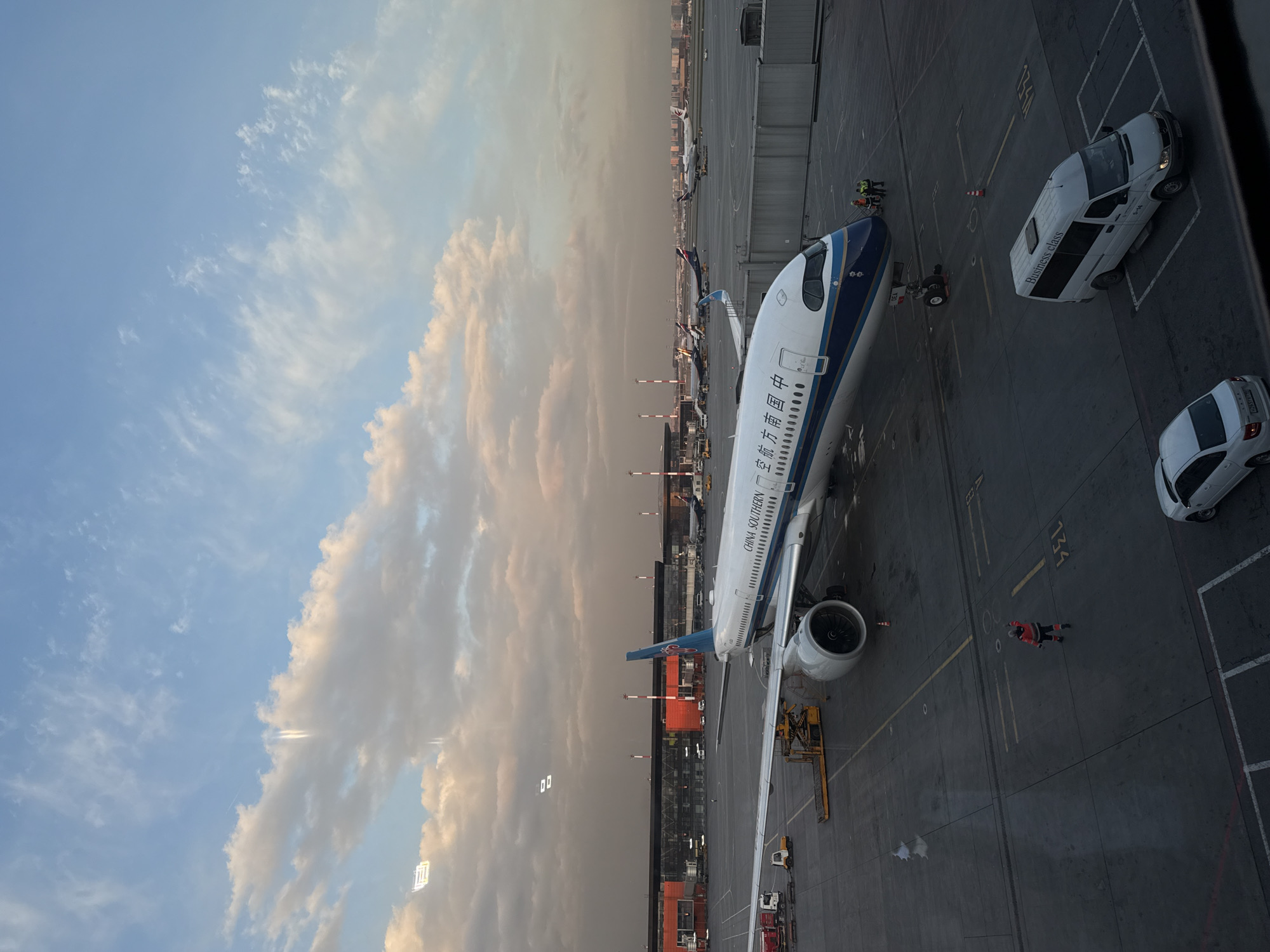 An aerial view from an airport terminal window captures a China Southern Airlines commercial aircraft parked on the tarmac. The image is oriented with the aircraft's nose pointing towards the left, set against a backdrop of a partly cloudy sky and distant airport infrastructure.
The dominant subject is a white and blue passenger jet, clearly identifiable by the "CHINA SOUTHERN" text and Chinese characters (中国南方航空) on its fuselage. The tail fin is blue, consistent with the airline's livery. One engine is prominent under the wing, and the aircraft's landing gear is visible.
Several airport ground crew members are present around the aircraft, some in high-visibility safety vests, indicating ongoing service or preparation. A white van, distinctly labeled "Business class" and "G-14", is parked near the aircraft's front section, suggesting passenger transport services. Another smaller white car is partially visible in the foreground. Ground support equipment, including a yellow vehicle, is positioned near the aircraft's wing. The tarmac features various yellow painted lines and numbers, such as "134", marking parking positions.
The sky above displays a blend of clear blue and soft, layered clouds, some catching a warm, golden light, indicative of late afternoon or early evening hours, possibly sunset. The horizon, stretching into the distance, reveals a hazy atmosphere and outlines of other parked aircraft and airport buildings. Given the context of Khimki, Russia, the scene is likely set at Sheremetyevo International Airport (SVO).