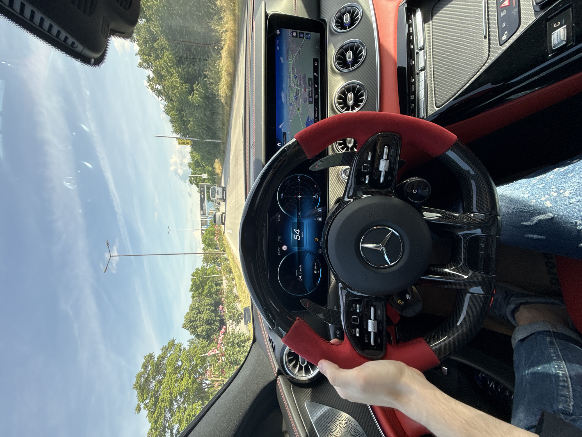 The image shows the interior of a Mercedes-Benz car, specifically the dashboard and steering wheel. The dashboard features a digital display showing a speed of 54 km/h and a navigation map. The navigation system is showing the location of Ulitsa Vasil Levski, in the city of Plovdiv, Bulgaria. A hand is visible on the steering wheel, and part of a person's leg in jeans is seen. The interior design includes red and black carbon fiber elements. Through the windshield, a road and trees can be seen, along with some street lamps and distant buildings. The sky is blue with some clouds.