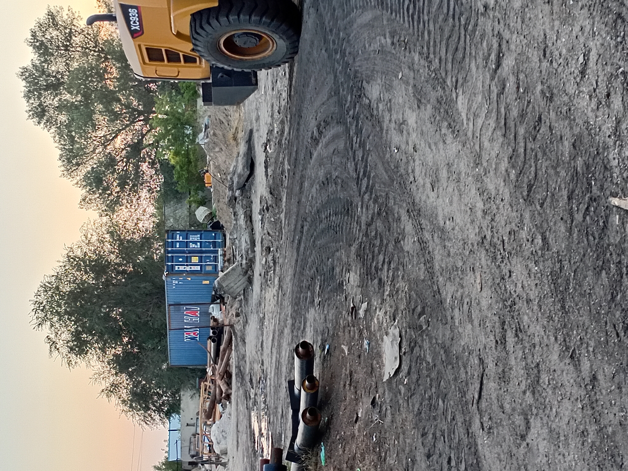 This image depicts an outdoor industrial or construction site, possibly a storage or processing area, in Semey, Kazakhstan. The scene is dominated by rough, dusty terrain with tire tracks visible. In the background, a large blue shipping container with "WAN HAI" branding is situated amongst piles of construction debris and materials, including pipes and what appears to be concrete rubble. To the right, the rear section of a yellow heavy machinery vehicle, possibly a loader, is visible, displaying the identification "XC936". The sky above is clear with a soft, orange hue, indicating either sunrise or sunset. Trees with dense foliage are scattered throughout the background, some illuminated by the low-angled sunlight. There are no people visible in the frame. The overall impression is one of an active, albeit perhaps unkempt, work zone.