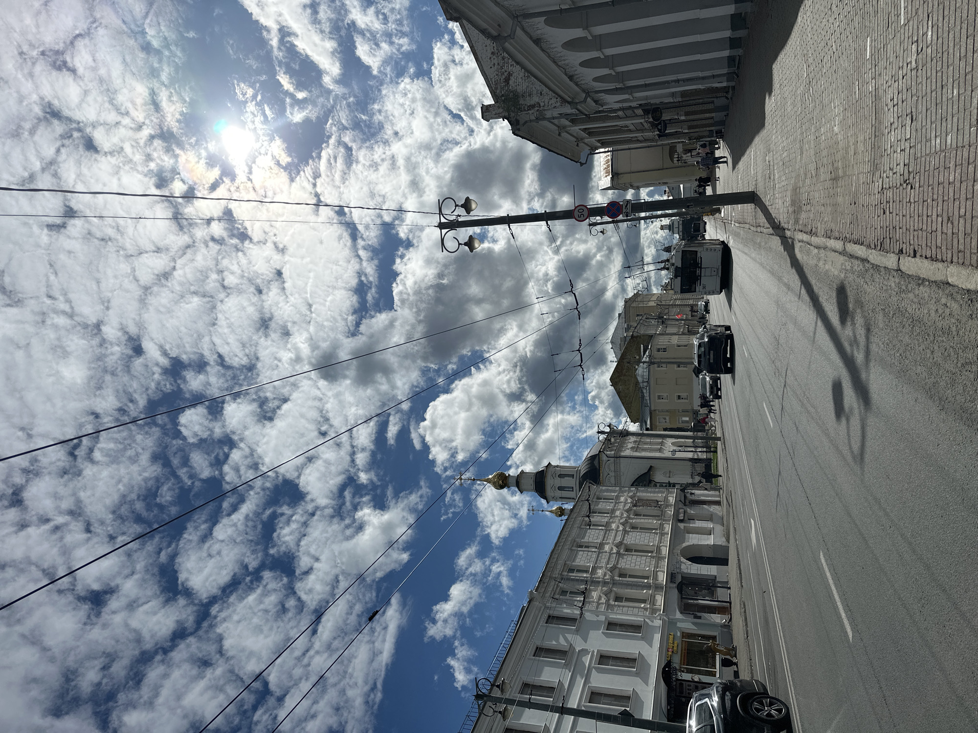 The image depicts a street scene in Vladimir, Russia, under a bright, partly cloudy sky. The sun is visible, casting a bright light and creating a rainbow effect through the clouds. The perspective is from the street level, looking up towards the sky and down the street lined with buildings.
The main subjects are the street itself, lined with buildings and with cars parked and driving on it. There are also overhead wires and streetlights visible. In the distance, a white building with an ornate facade and a golden-domed church spire can be seen. People are present, walking on the sidewalk and in the street. A trolleybus is also visible, as are several cars.
The scene appears to be set during the daytime, likely late morning or early afternoon, given the brightness of the sun and the shadows cast. The weather is pleasant, with a mix of bright sunshine and scattered clouds. The architecture of the buildings suggests a historical urban setting, consistent with the specified location of Vladimir, Russia.
Notable details include the prominent overhead wires, characteristic of electric transport in many Russian cities. The cobblestone sidewalk in the foreground on the right side of the street is also a distinct feature. There is some visible text on the buildings, although it is not clear enough to be fully deciphered. One sign on a lamppost reads "50," likely indicating a speed limit. The overall mood is one of a typical urban day, with everyday activities taking place.