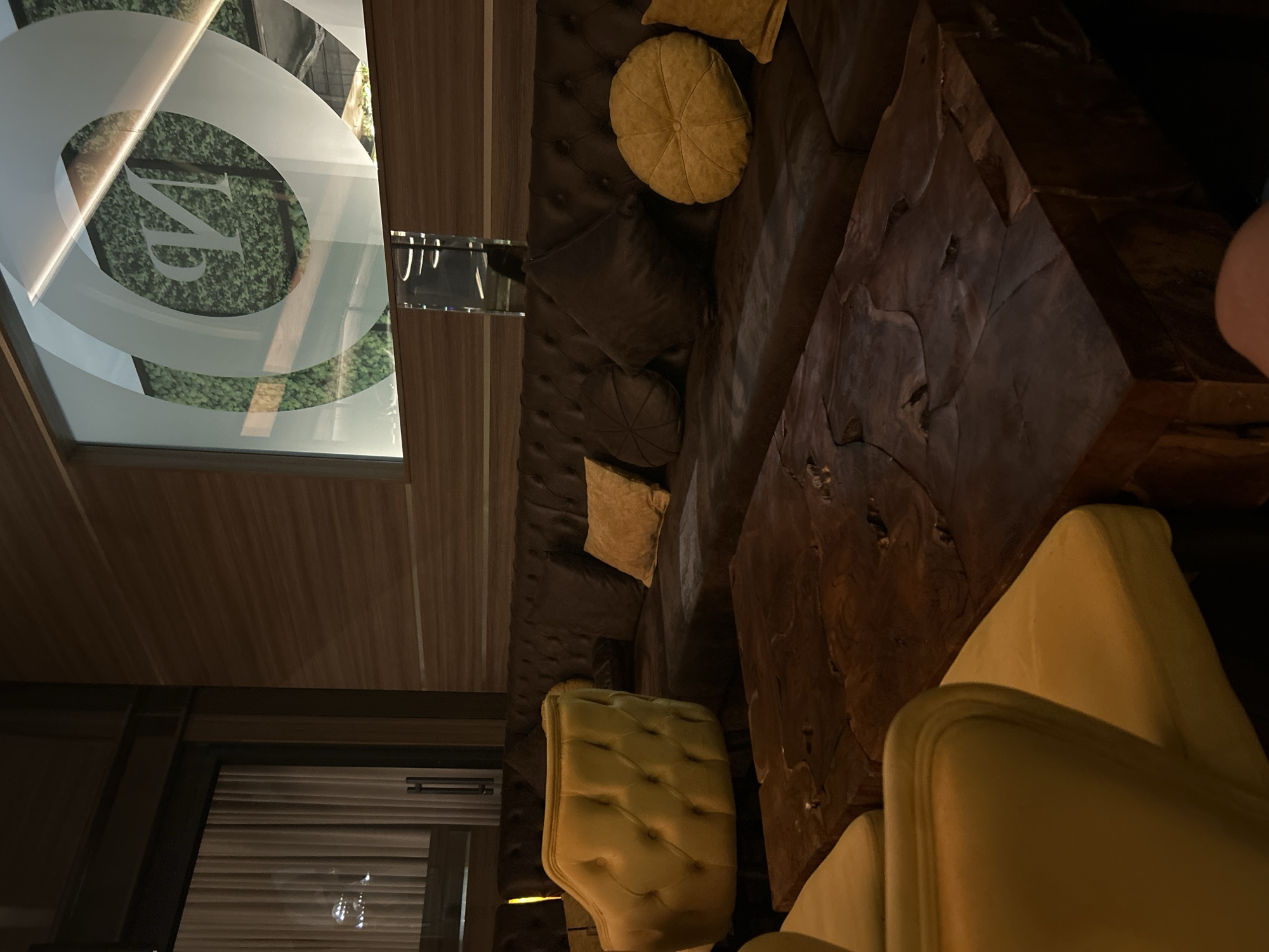 This image depicts an interior scene, likely a lounge or bar area. The foreground is dominated by a large, dark wood coffee table with a rough, naturalistic surface. To the left, a plush, tufted yellow armchair is partially visible. Behind the coffee table, a long, dark brown tufted sofa stretches across the frame, adorned with several decorative pillows in various shades of brown and yellow.
In the background, a large window or glass partition features a prominent white logo, a stylized circular design with a "V" or "N" shape within it. Behind this logo, a lush green wall covering, possibly artificial foliage, is visible. The walls of the establishment appear to be paneled with dark wood. To the far left, a window with sheer curtains is visible. The lighting is dim and ambient, creating a cozy and sophisticated atmosphere. The image does not provide any visual clues to indicate its specific location in Burgas, Bulgaria.