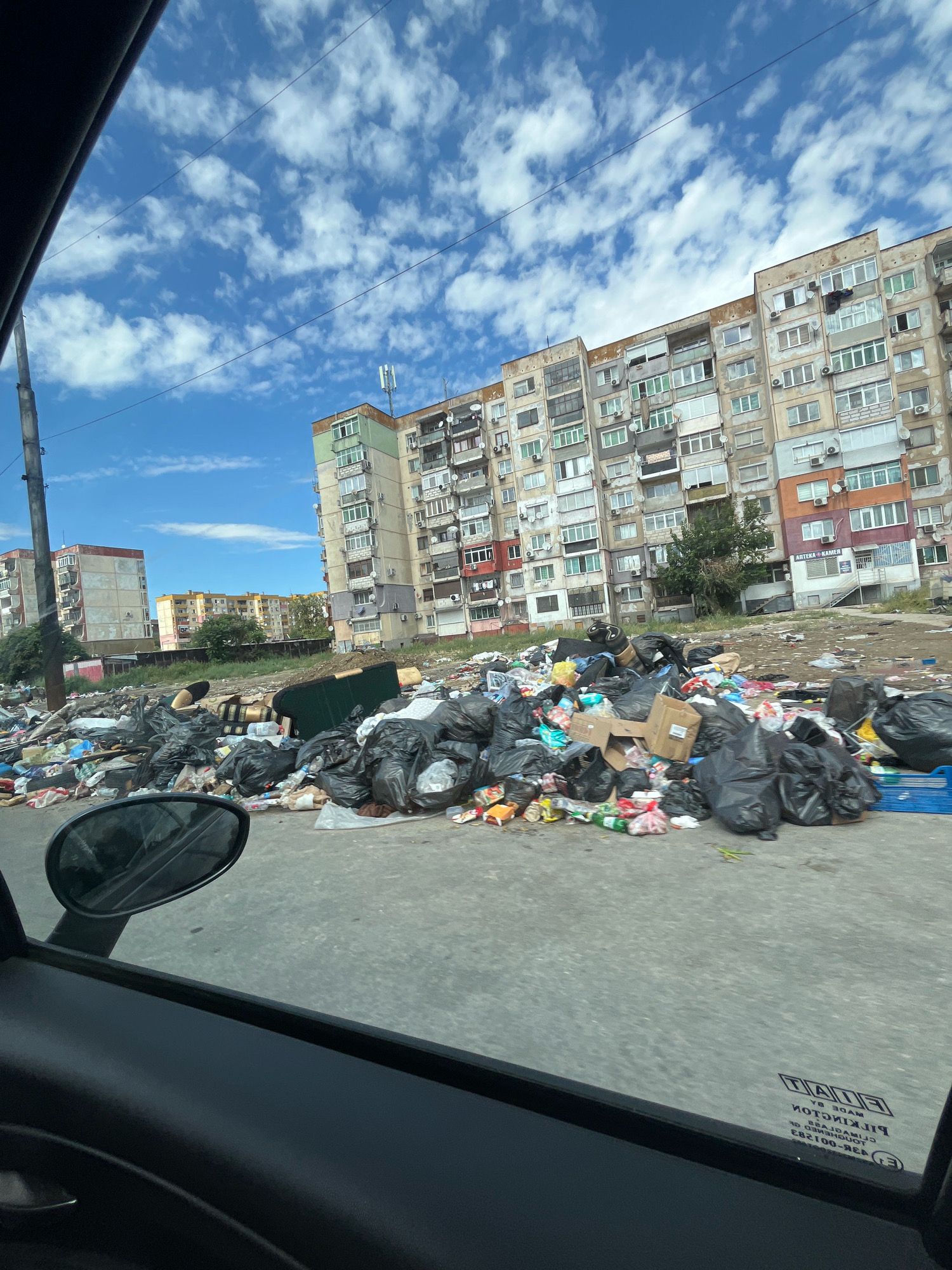 The image depicts a scene of urban neglect and overflowing refuse in Plovdiv, Bulgaria. A significant accumulation of garbage bags, discarded items, and debris is piled high on a paved surface, obscuring much of the ground. In the background, a row of multi-story apartment buildings stands, some appearing weathered and in disrepair, with visible satellite dishes and air conditioning units on their facades. A few trees are scattered between the buildings and the garbage. A utility pole is visible on the left side of the frame. The sky above is partly cloudy with a bright blue expanse. The perspective is from inside a vehicle, with the car's side mirror and door frame visible in the foreground. The overall impression is one of an unkempt and unsanitary urban environment.