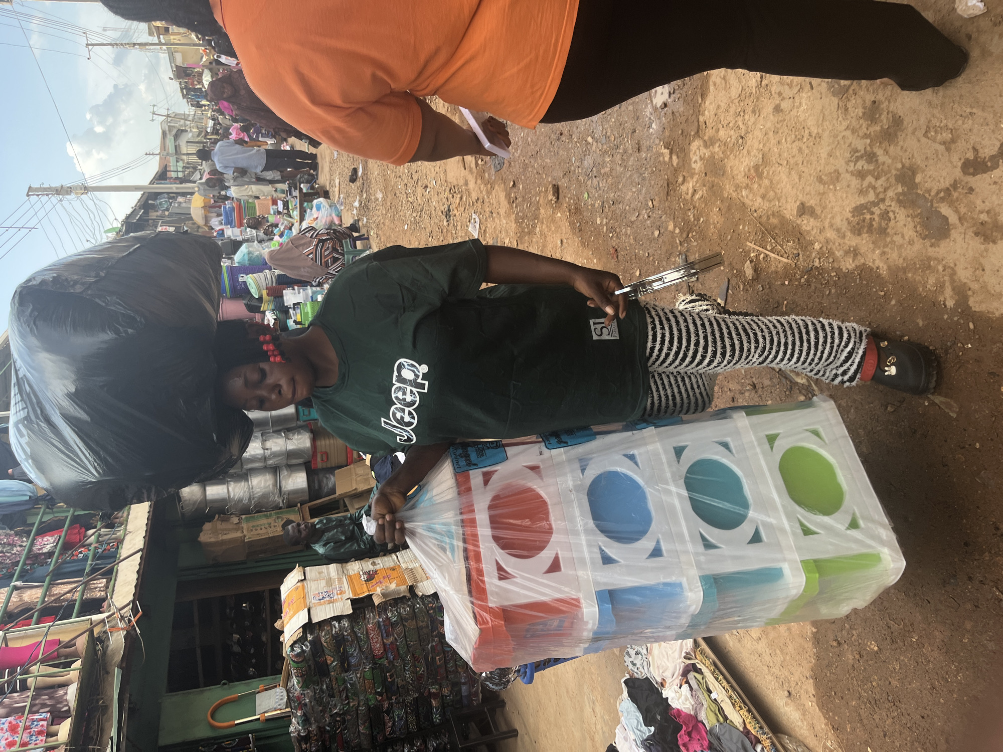 In Jos, Nigeria, a woman carries a large black trash bag balanced on her head, her face partially obscured by the bag. She wears a dark green t-shirt with a white "Jeep" logo and striped pants. In her right hand, she holds a clear plastic bag filled with a stack of colorful plastic storage drawers. The drawers are red, blue, and green, with white frames. She also has a metal object, possibly a stapler, in her hand. The scene is a bustling outdoor market, with stalls selling various goods, including fabrics, cookware, and plastic items. Other people are visible in the background, suggesting a busy commercial area. The ground is unpaved and dusty, typical of an open-air market.