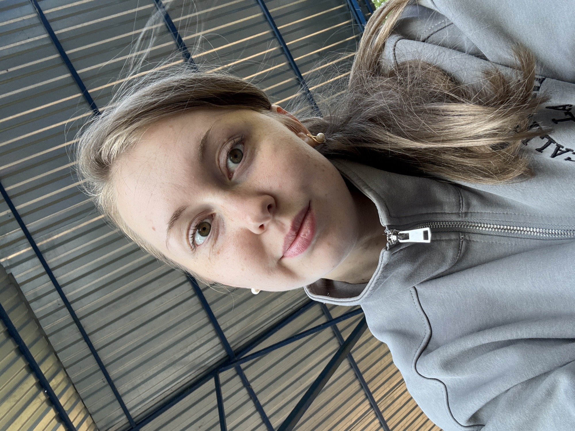 The image captures a selfie of a young woman taken from a low, upward-angled perspective. She has a fair complexion with faint freckles on her nose and cheeks, greenish-brown (hazel) eyes, and light brown or ashy blonde hair that falls loosely around her face and over her shoulders. She wears a small, gold-toned hoop earring with a white pearl in her right ear. Her expression is calm, with a slight, subtle smile, as she looks directly at the camera.
She is dressed in a light gray or beige-gray zippered jacket or sweatshirt, featuring a prominent silver-toned zipper. On the right side of her chest, dark embroidered text is partially visible, with the letters "CAL" clearly discernible.
The background consists of an overhead view of a roof or ceiling structure, characterized by corrugated metal panels in shades of silver or gray, intersected by dark blue or black structural beams. The arrangement forms a distinct linear pattern. The scene is illuminated by bright natural daylight, suggesting an outdoor or semi-enclosed setting during daytime.