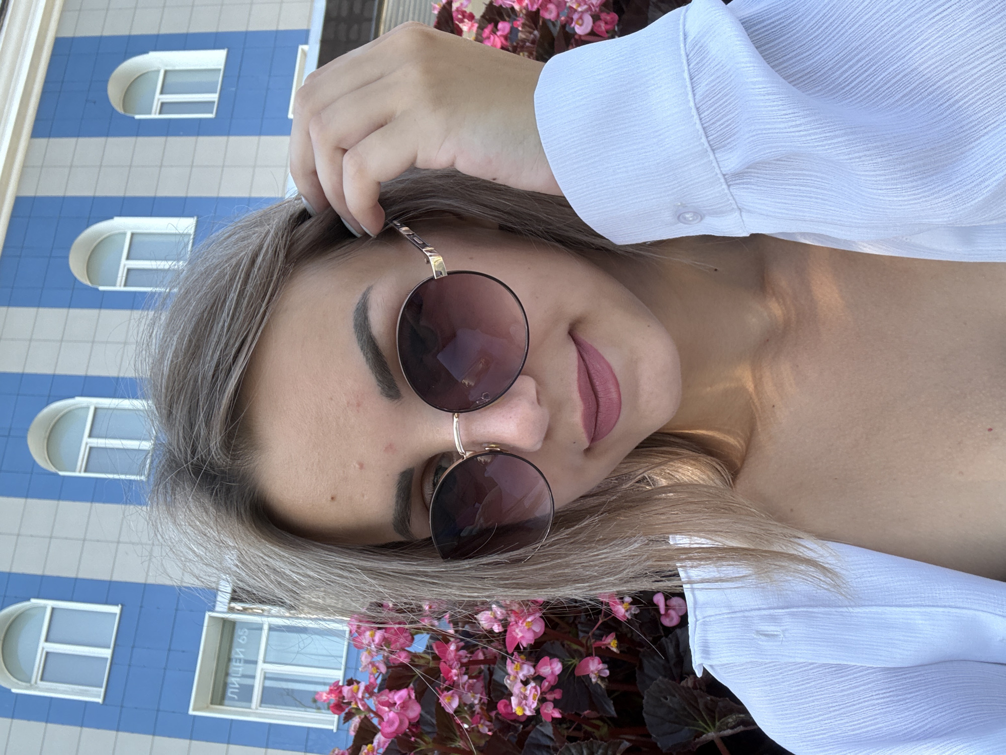 A young woman with blonde hair and sunglasses is taking a selfie outdoors. She is wearing a white shirt and has her hand on her hair. Behind her, there is a building with blue and white vertical stripes and arched windows. In the foreground, there are pink flowers. The building appears to be a school or educational institution, as one window has the text "ЛИЦЕЙ 65" (Lyceum 65) visible. This detail suggests the location is Lyceum No. 65 in Yuzhno-Sakhalinsk, Russia.
