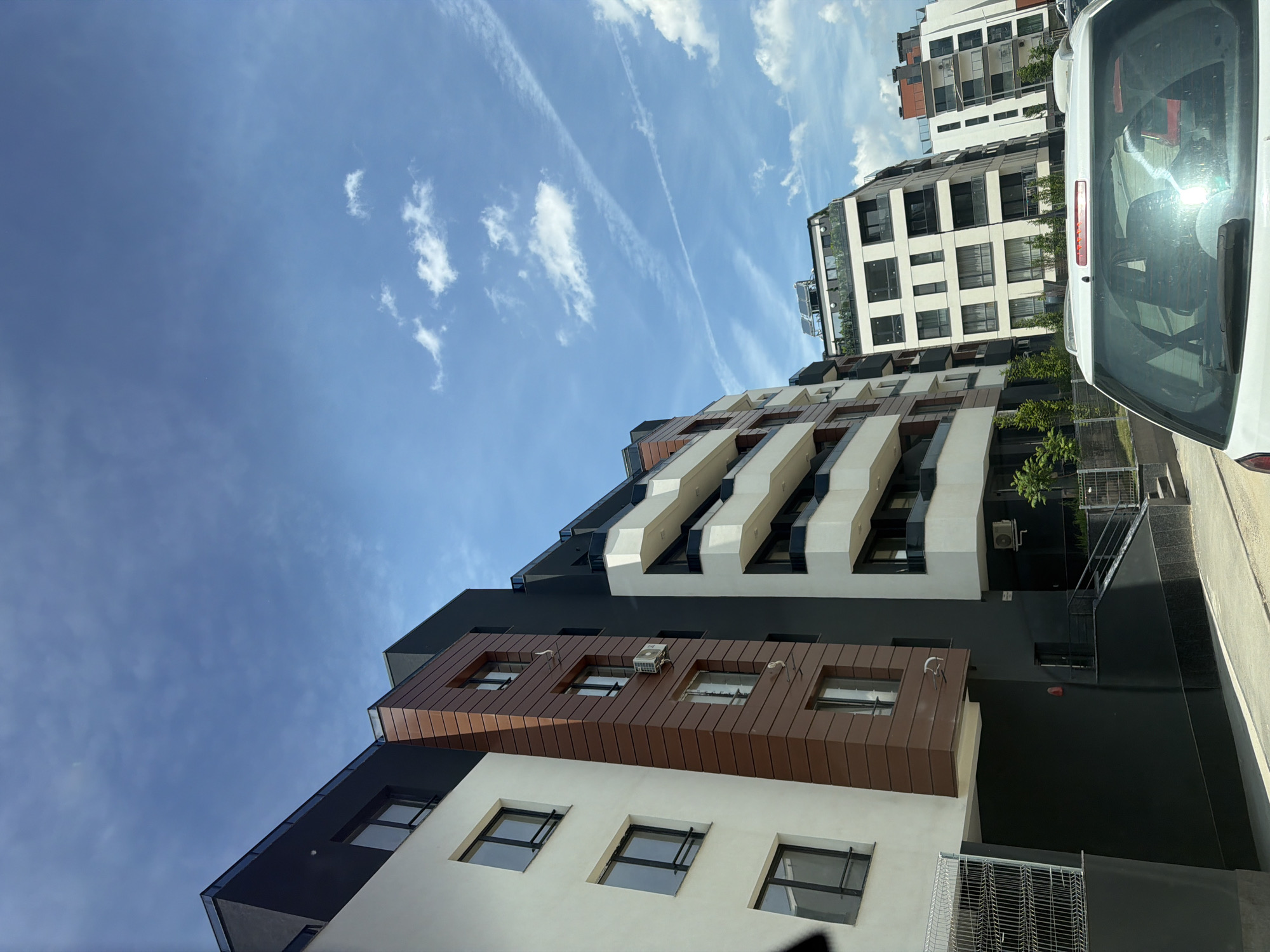 The image displays an urban street scene in Sofia, Bulgaria, rotated 90 degrees clockwise. It features modern apartment buildings under a bright blue sky, with the rear of a white car partially visible on the right.
The central and left portions of the image are dominated by multi-story residential buildings with contemporary architectural designs. These buildings showcase a facade composed of contrasting elements: dark grey and black sections, clean white walls, and warm brown-toned paneling, some with vertical slats. Rectangular windows with dark frames are systematically arranged across the buildings. Several exterior air conditioning units are mounted on the walls. One building features distinctive projecting, angular balcony structures in white and black.
On the far right, the back of a white car, likely a hatchback or SUV, is seen parked. Its tinted rear window reflects the bright sky and surrounding structures.
The upper part of the frame is filled with a clear, vivid blue sky, dotted with scattered white clouds. Several distinct, linear contrails (chemtrails) stretch diagonally across the sky, indicating overhead air traffic. The scene is illuminated by bright daylight, suggested by the clear sky and lack of deep shadows, implying sunny weather. No people or specific activities are visible. No discernible text is present on the buildings or car.