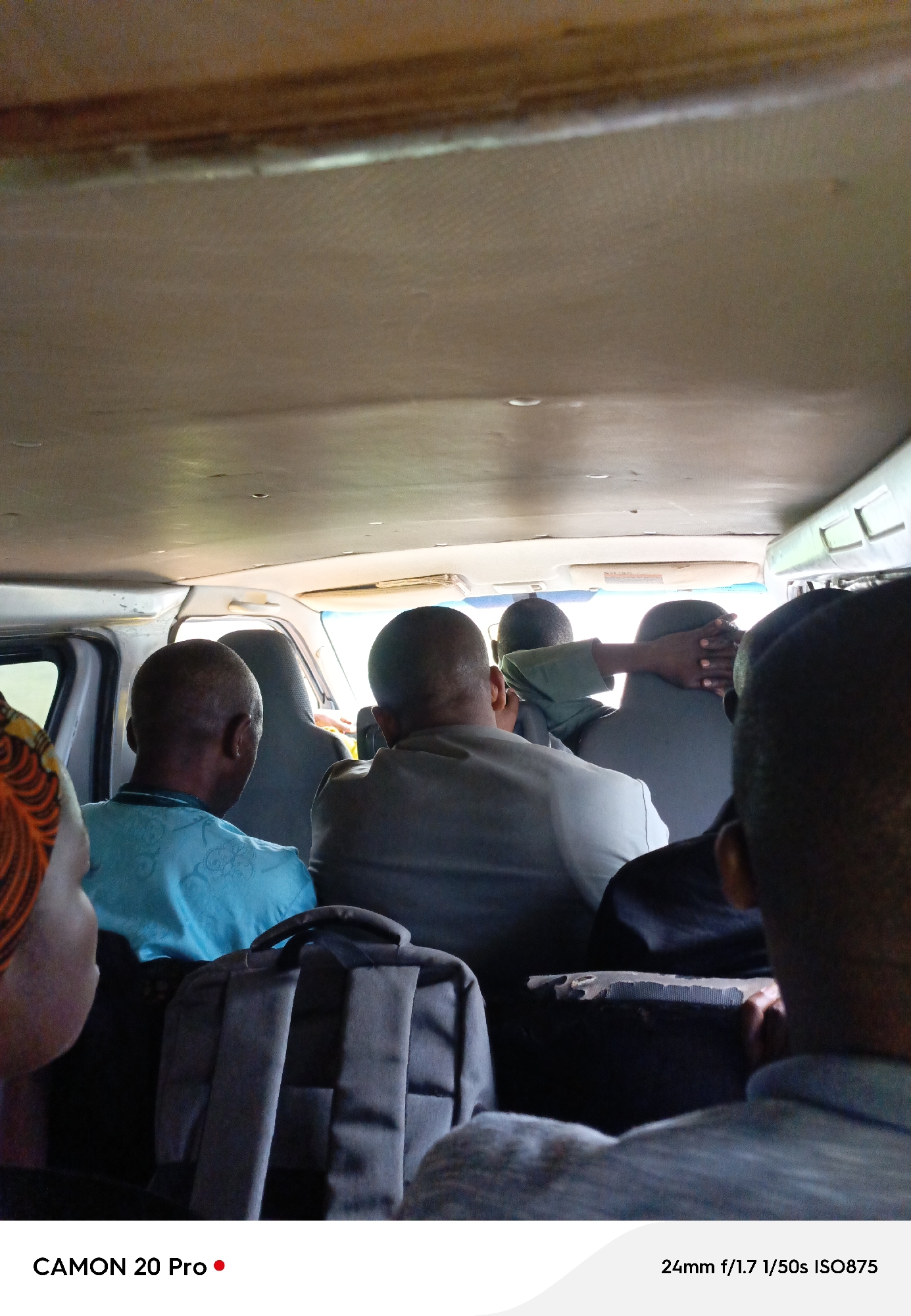 The image captures the interior of a van in Nguroje, Nigeria, with several passengers seated. The focus is on the backs of their heads and upper bodies, suggesting a forward-facing perspective from within the vehicle. The van's ceiling is visible, appearing to be made of a light-colored, textured material, with some signs of wear and tear. Natural light filters in from the windows, creating soft illumination. In the foreground, on the left, a person wearing a colorful headscarf is partially visible. A dark backpack rests on the seat in the middle of the frame. The passengers are dressed in casual attire, and their postures indicate they are traveling. The overall scene suggests a moment of travel within a community setting.