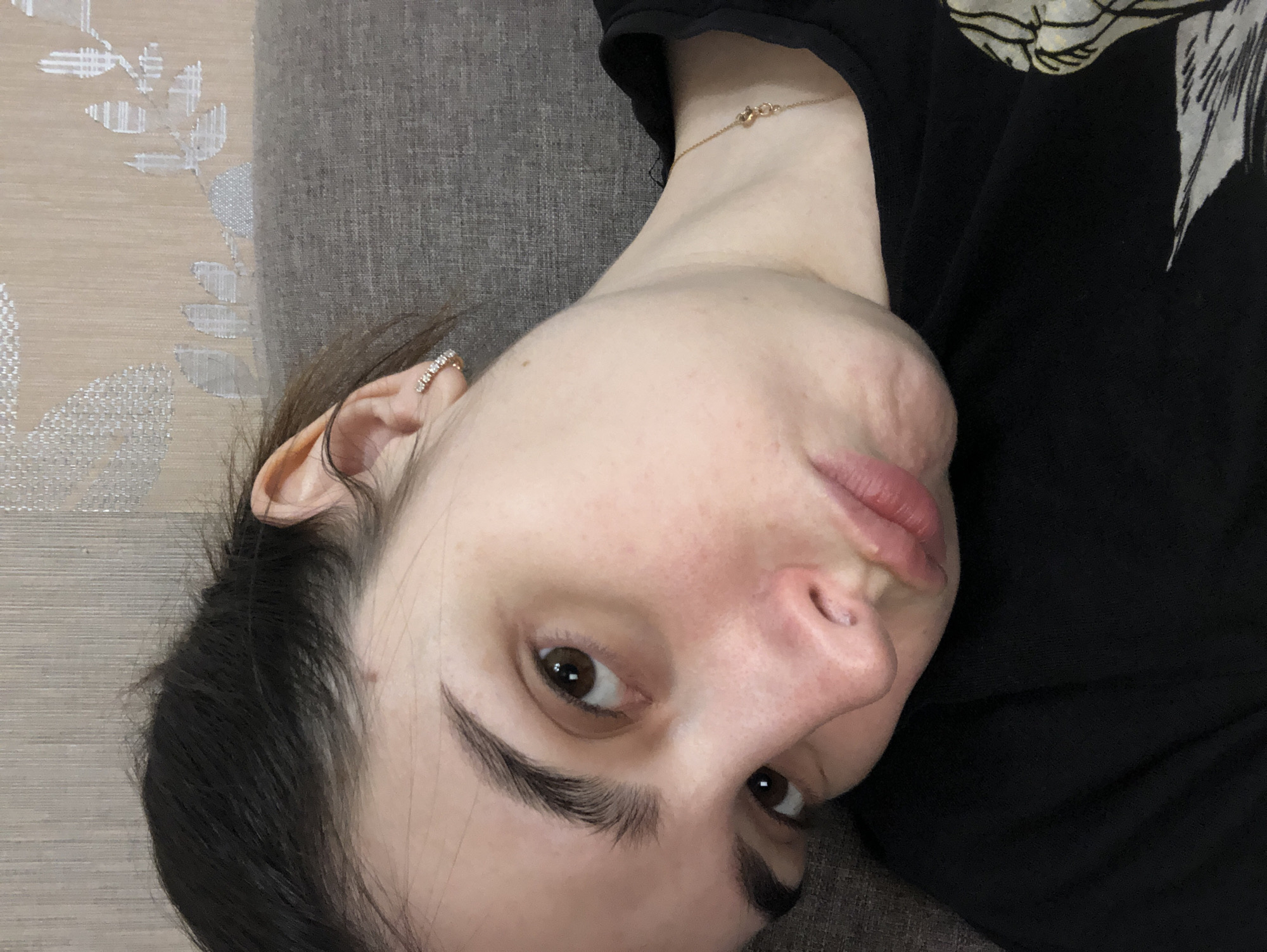 A close-up, rotated image shows a light-skinned woman with dark hair lying on a gray-brown textured surface, possibly a couch or cushion. She wears a black top, and a delicate gold chain is visible around her neck. The woman has dark eyebrows and brown eyes and is wearing an earring with small stones on her left ear. A patterned wall is partially visible in the upper left corner of the image. There is no verifiable information to suggest the location is in the city of Chaikovsky, Russia.