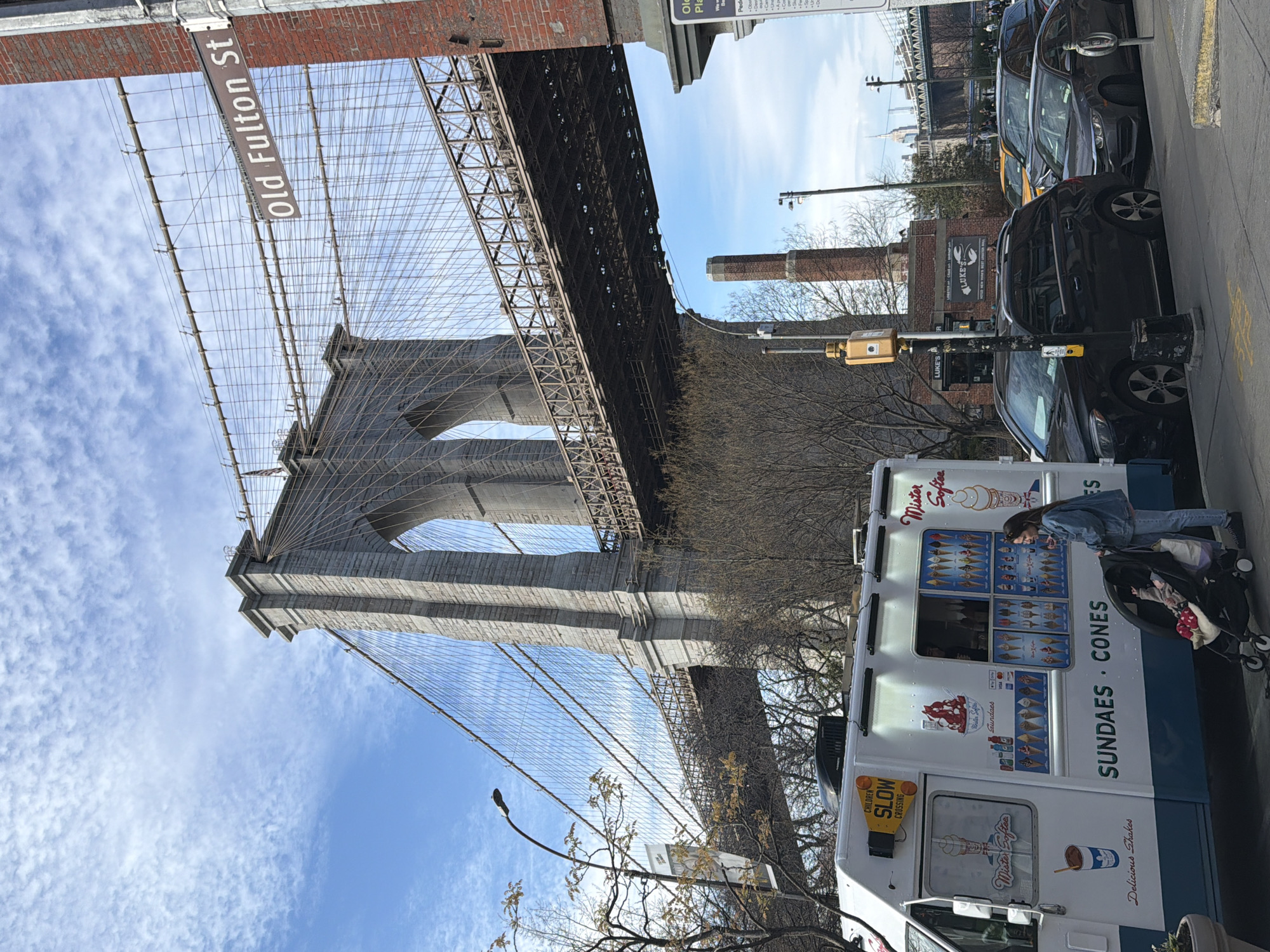This is a street-level view of the Brooklyn Bridge in New York City. The bridge's massive stone tower looms overhead, with "Old Fulton St" sign. A Mister Softee ice cream truck is parked on the side of the road. A woman is by the truck, possibly buying ice cream. Cars are parked along the street as well. The sky is bright blue with scattered clouds. A sign for Luke's Lobster is visible on a brick building.