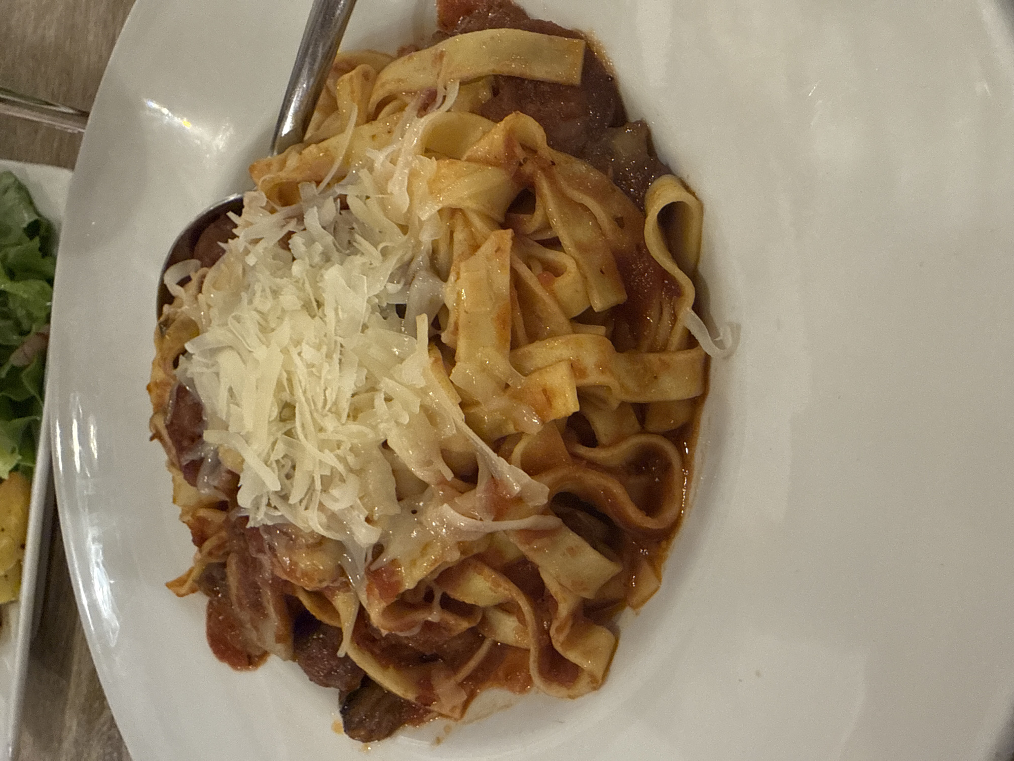A close-up, high-angle shot captures a serving of pasta on a white, oval-shaped plate, likely in an indoor dining setting in Varna, Bulgaria. The main dish features broad, flat pasta noodles, possibly fettuccine or tagliatelle, coated in a rich, reddish-brown sauce that appears to contain pieces of meat or mushrooms. A generous amount of grated white cheese, resembling Parmesan, is piled on top of the pasta, partially melted and clinging to the noodles. A metal spoon is partially submerged in the food on the left side of the plate.
In the blurred background to the left, a portion of another white plate is visible, containing a side dish of green salad, suggesting a meal with multiple components. The lighting is artificial, casting subtle reflections on the shiny surface of the plate. The focus is entirely on the food, presenting a delicious and ready-to-eat meal. No people or text are visible in the image.