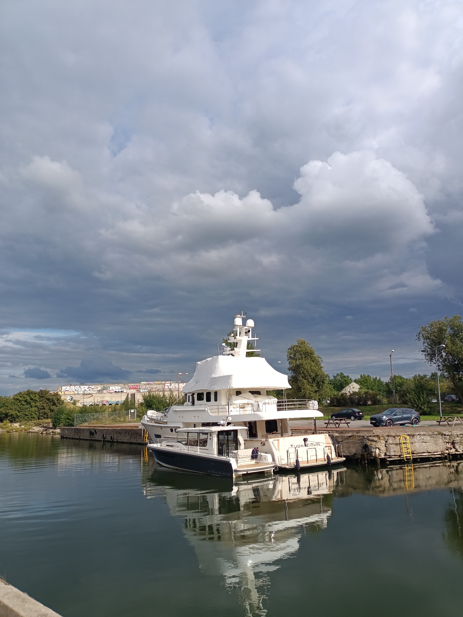 A large, white motor yacht is docked next to a smaller, dark-hulled boat in a harbor. The yacht has multiple decks and appears to be covered in protective sheeting on its upper sections. The smaller boat has the name "NIMBUS" visible on its stern. Two individuals are visible on the deck of the smaller boat. In the background, a concrete dock runs along the waterfront, with trees and buildings beyond. Two cars are parked on a paved area behind the dock. The sky is filled with dramatic, dark clouds, suggesting an impending storm. The water is calm and reflects the sky and the boats. This scene is located in Tallinn, Estonia, likely in a port or marina area.