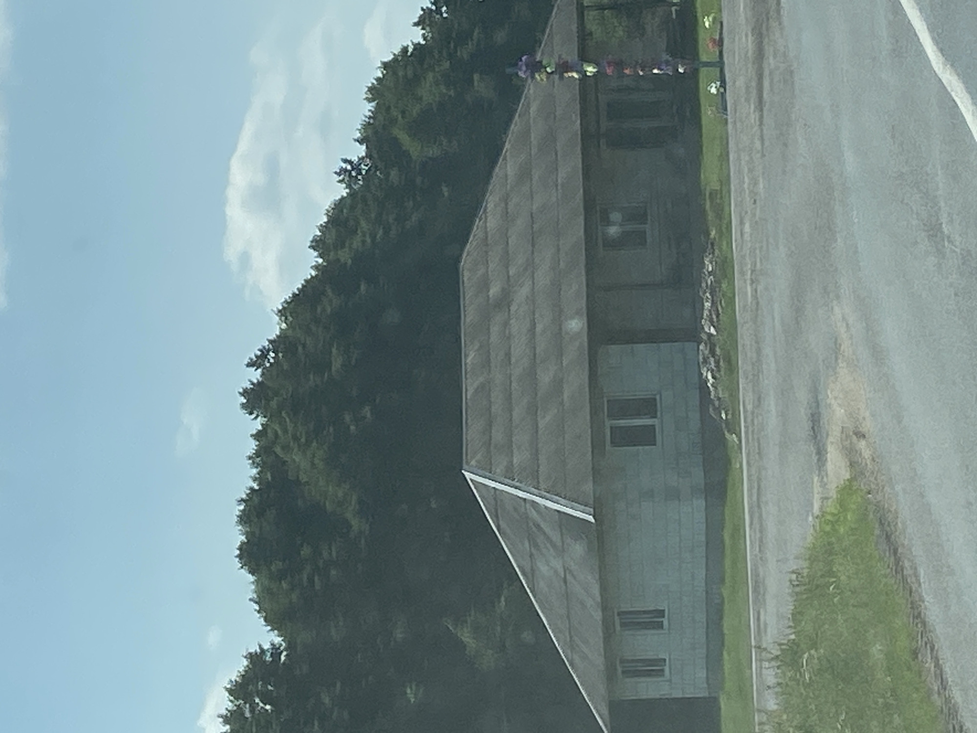 The image depicts a rural scene in the village of Volchinsky, within the Volchinsky rural council, Belarus. In the foreground, a paved road with a white line marker is visible. To the left of the road, a grassy area with weeds and a dirt patch is present. The main subject is a single-story house with a gray, corrugated roof and light-colored cinder block walls. The house has several windows. To the right of the house, a pole adorned with colorful decorations, possibly flowers or ribbons, stands near the roadside. A small pile of rocks is located near the foundation of the house. Behind the house, a dense line of dark green trees forms a backdrop against a light blue sky with scattered clouds. The overall setting appears to be a sunlit day in a countryside location.