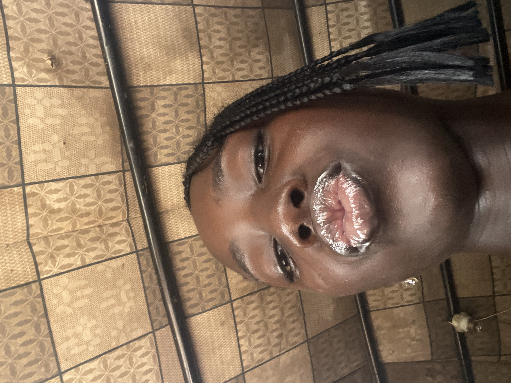 A low-angle, medium close-up shot shows a young Black person with braids and metallic lipstick, looking directly at the camera with their lips puckered. The background features a patterned ceiling made of square tiles in beige and brown, with a dark horizontal bar running across the upper portion of the frame. A small light fixture is visible on the left side of the frame, hanging from a wire. The image is likely taken in a residential setting within Galadimawa, Nigeria.