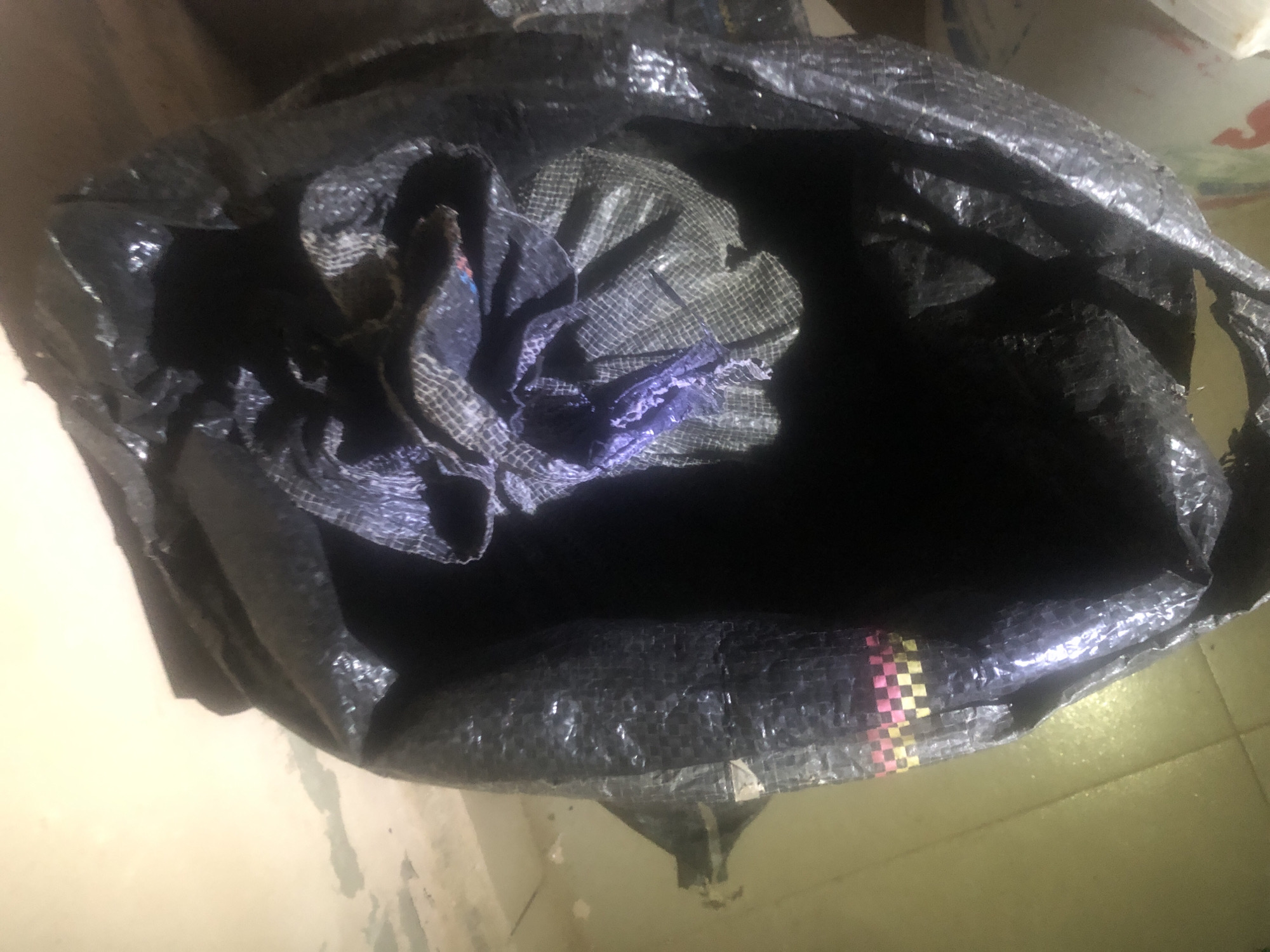 The image shows an open black plastic bag. The bag is partially filled with a dark substance, and appears to have some crumpled fabric or material inside, some of which is purple and checkered. The bag is sitting on a floor that appears to be tiled. The background is out of focus but appears to be a wall and possibly a wooden surface. The image is taken from a high angle, looking down into the bag. There are no people or animals visible in the image. The location in the city of Wukari, Nigeria is not discernible from the image.