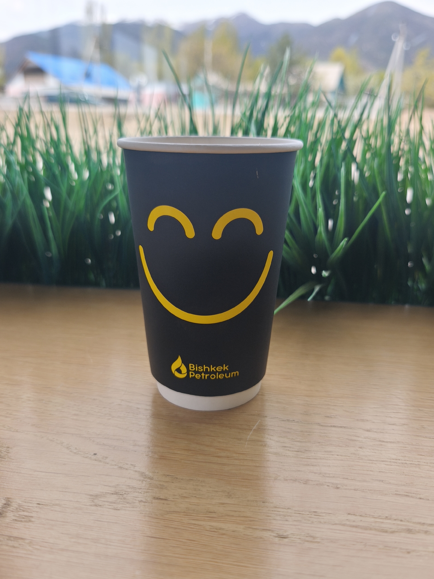A close-up shot shows a black paper cup with a yellow smiley face and the words "Bishkek Petroleum" and a logo printed on it. The cup sits on a light brown wooden surface, possibly a table. In the blurred background, there's a view of green grass-like plants, buildings with a blue roof, and mountains under a slightly overcast sky. The image seems to be taken in an outdoor setting, most likely in the city of Bishkek, Kyrgyzstan, based on the branding on the cup.