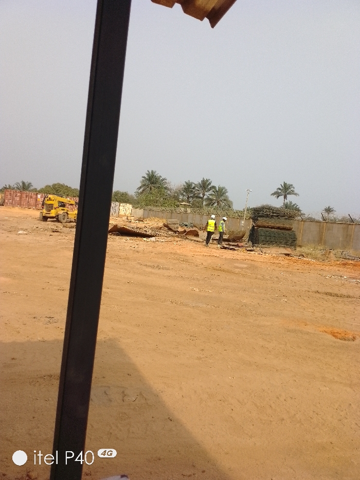The image depicts an outdoor industrial or storage yard in Hoche, Nigeria, under a light, hazy sky. A dark, vertical structural column partially obstructs the left foreground, with a corrugated roof edge visible at the top. The ground is unpaved, reddish-brown dirt, covering most of the lower half of the frame.
In the midground, two individuals are standing on the dirt. Both wear yellow-green high-visibility vests and white hard hats. They are positioned near a concrete perimeter wall that extends horizontally across the scene, topped with what appears to be barbed or razor wire. To the immediate right of the individuals, a large stack of bundled metal mesh or rebar is present. To their left, scattered debris, including what resembles wood and metal fragments, lies on the ground. Further to the left in the midground, a yellow mobile crane truck with an extendable boom is parked. Behind the crane truck, several stacked red-brown shipping containers are visible. A light pole with a fixture is situated behind the individuals, near the concrete wall.
The background features numerous palm trees and other green vegetation behind the wall and containers. The "itel P40 4G" watermark is present in the lower-left corner. No direct interaction between the individuals or specific actions are clearly discernible; they appear to be observing or standing within the yard.