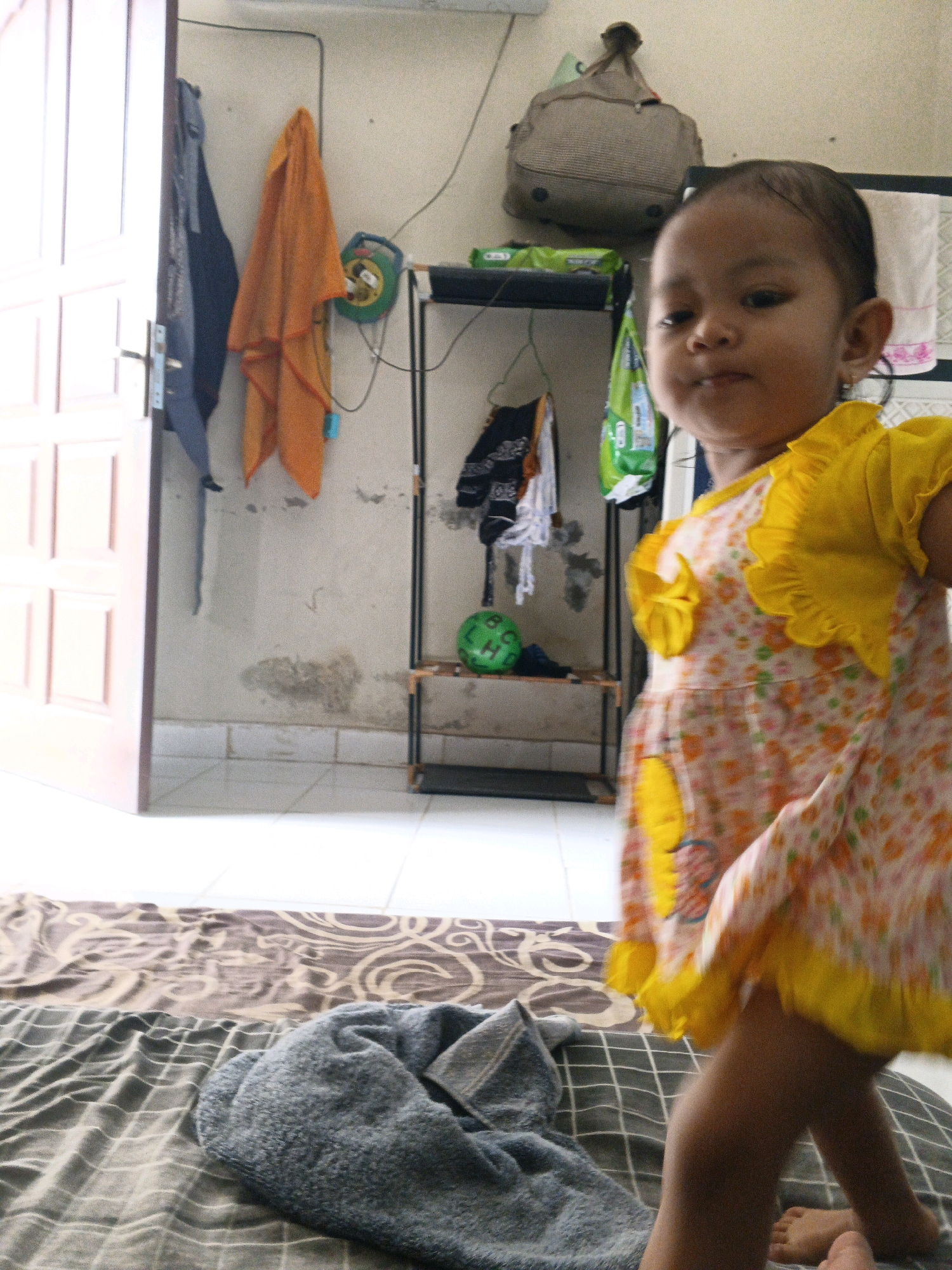 An outdoor, sunlit scene captures a young toddler girl with dark hair and brown eyes standing in what appears to be an open-air laundry area or a makeshift clothing storage space. She is wearing a light-colored dress with floral patterns of pink and orange, accented by bright yellow ruffled sleeves and a yellow flower appliqué on the chest. To her left, a light pink door is ajar, revealing its paneling and hardware. Behind her, a rack of various items of clothing is hung, including a dark blue garment, an orange cloth, and other unidentifiable fabrics. A green ball sits on the bottom shelf of the rack. The wall behind the rack is textured and shows signs of age and possible water damage, with patches of discoloration. A woven bag hangs from the wall above the rack. The floor in the foreground consists of patterned blankets, one with a brown swirling design and another with a grey checkered pattern. A grey, folded towel or blanket rests on the checkered blanket. The scene is likely in Denpasar, Indonesia, a region known for its tropical climate and open-air living spaces.