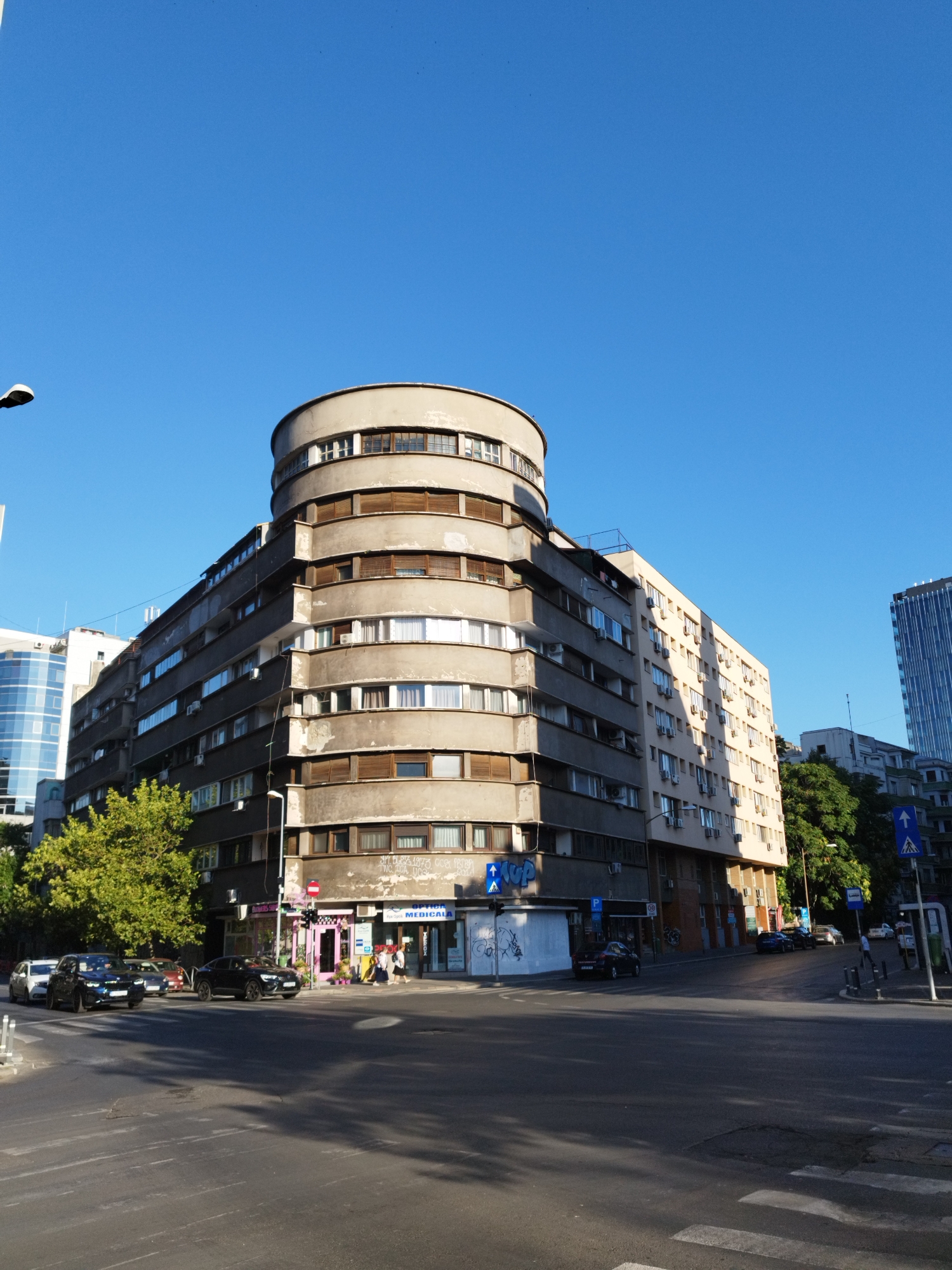 This image depicts a street scene in Bucharest, Romania, focusing on a distinctive curved, Brutalist-style building at a street intersection. The building, constructed with grey concrete, features multiple stories with numerous windows and balconies. A cylindrical section protrudes from the top of the building, adding to its unique architectural character. The lower levels of the building house commercial establishments, including shops with bright signage, and a visible sign for "OPTICA MEDICALA".
Several cars are present on the street, some parked and others in motion. Pedestrians are visible on the sidewalks and crossing the street, indicating a moderately busy urban environment. The sky is clear and blue, suggesting a sunny day. Trees line the street in some areas, providing greenery against the concrete and glass of the urban landscape. The presence of streetlights, traffic signs, and crosswalks further defines the setting as a typical city intersection. In the background, other buildings of varying architectural styles, including a modern high-rise with a glass facade, are visible. The overall impression is one of a dynamic urban environment characterized by a mix of historical and contemporary architecture.