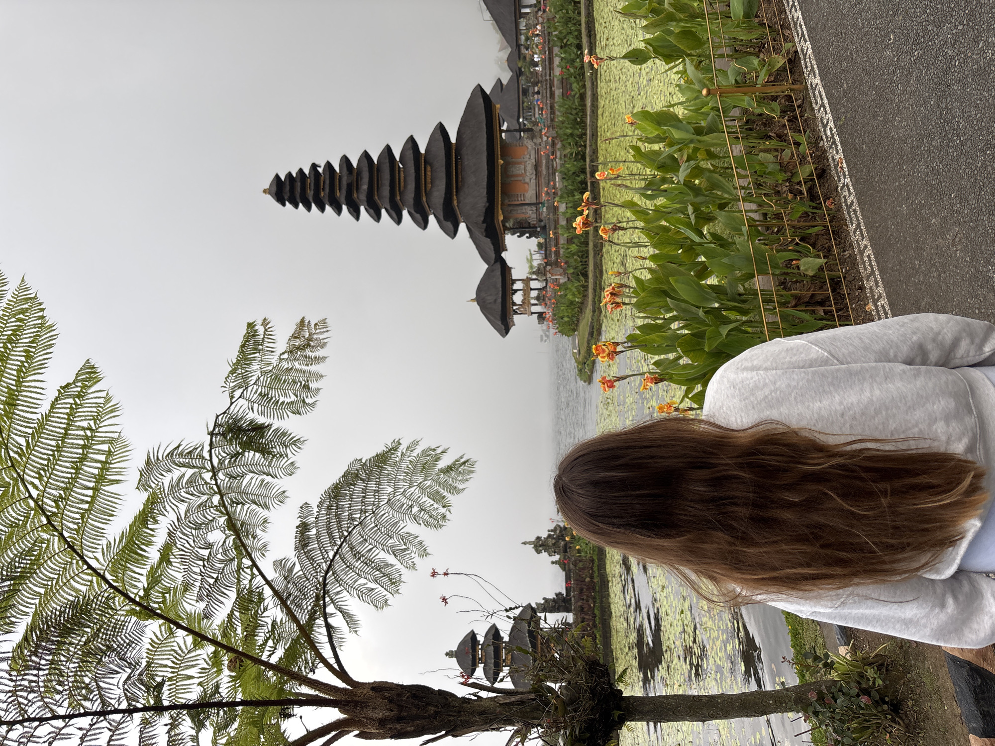 A low-angle, landscape orientation photograph shows a woman facing away from the camera and looking toward a large pagoda-style structure, located in Baturiti, Indonesia. The woman, cropped from the upper-mid torso upward, wears a light gray sweatshirt and has long, wavy brown hair. In the foreground, to the left, is a fern tree. The pagoda is composed of several layers that decrease in size as they ascend. Between the woman and the pagoda, there are yellow and orange flowering plants. The ground consists of gray pavement with a decorative white border. The sky is overcast and light gray. Water is visible in the background around the pagoda and the immediate foreground, suggesting the setting is near a lake or pond.