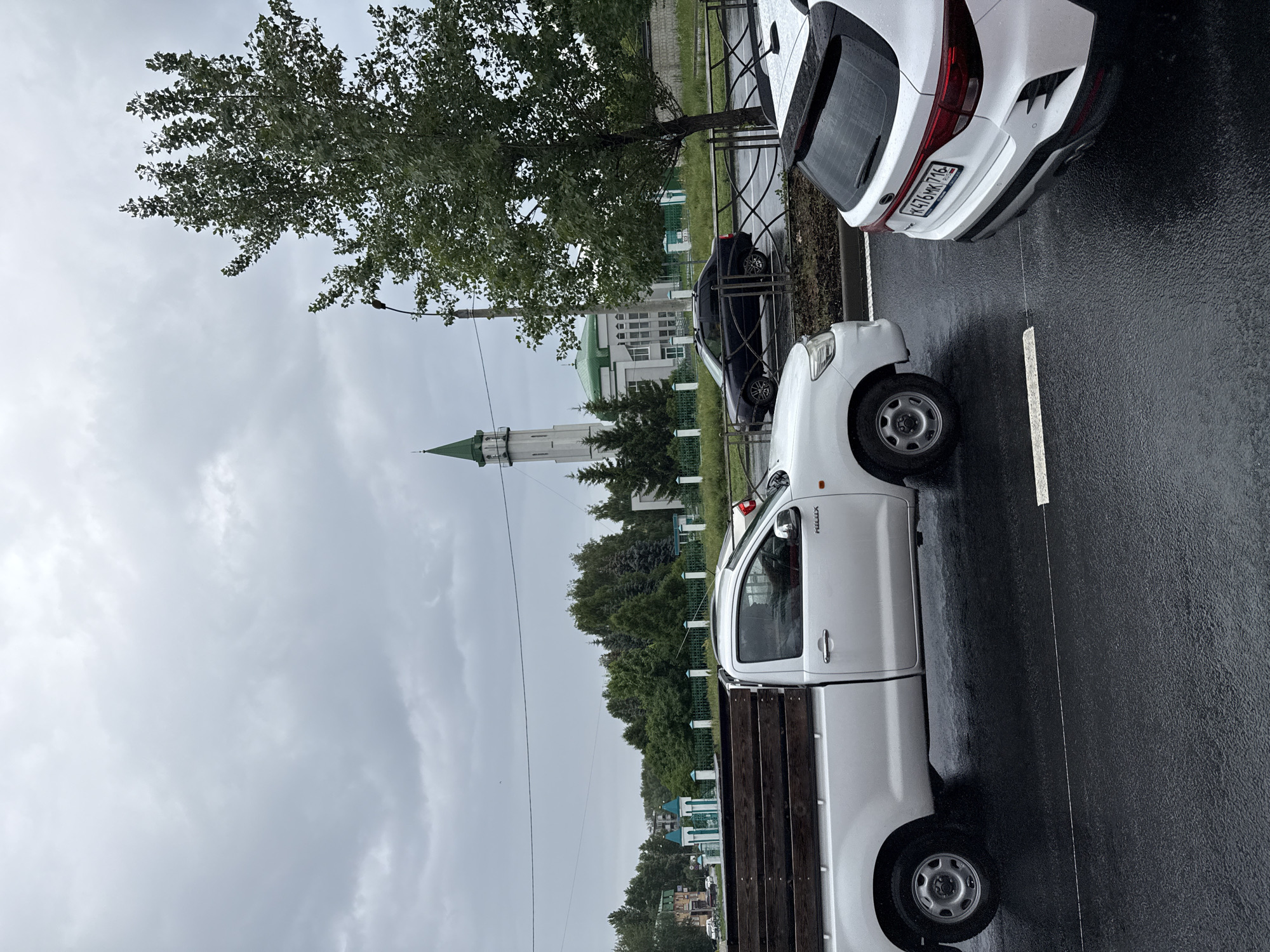 In Kazan, Russia, on a cloudy day with wet pavement, a white pickup truck with a wooden bed is parked on the right side of the street. A dark sedan is visible behind it. In the background, a white mosque with a green dome and minaret stands behind a green fence. A large tree with green leaves is on the right side of the frame. Another white SUV is parked in the foreground on the right.