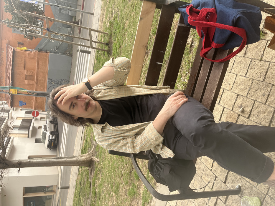 The image captures a male individual seated on a wooden park bench with black metal supports, located in Tel Aviv-Yafo, Israel. The man, appearing in his 20s or 30s, has medium-length dark wavy hair and a mustache. He is wearing a light-colored plaid button-up shirt unbuttoned over a black t-shirt, dark trousers cuffed at the ankles, and a wristwatch with a dark strap on his left wrist. His right hand is placed on his forehead, and his gaze is directed at the camera.
A black drawstring bag rests on the bench to his left, and a dark blue bag with red straps is on the bench to his right. A translucent green cup with a lid and straw is visible on the light-colored paved ground to the lower left of the bench. Beyond the paved area, a strip of grass precedes a street with a white crosswalk. A white pickup truck is parked on the street. Buildings line the street, including a light-colored building with balconies and visible Hebrew text on a ground-level window, and a reddish-orange building with external stairs. A "No Entry" sign and a yellow pole with a blue sign indicating "30" in a red circle are visible on the street, confirming the location as consistent with Israeli signage. A tree with a bare trunk is situated between the grass strip and the street. The sky appears overcast or hazy.