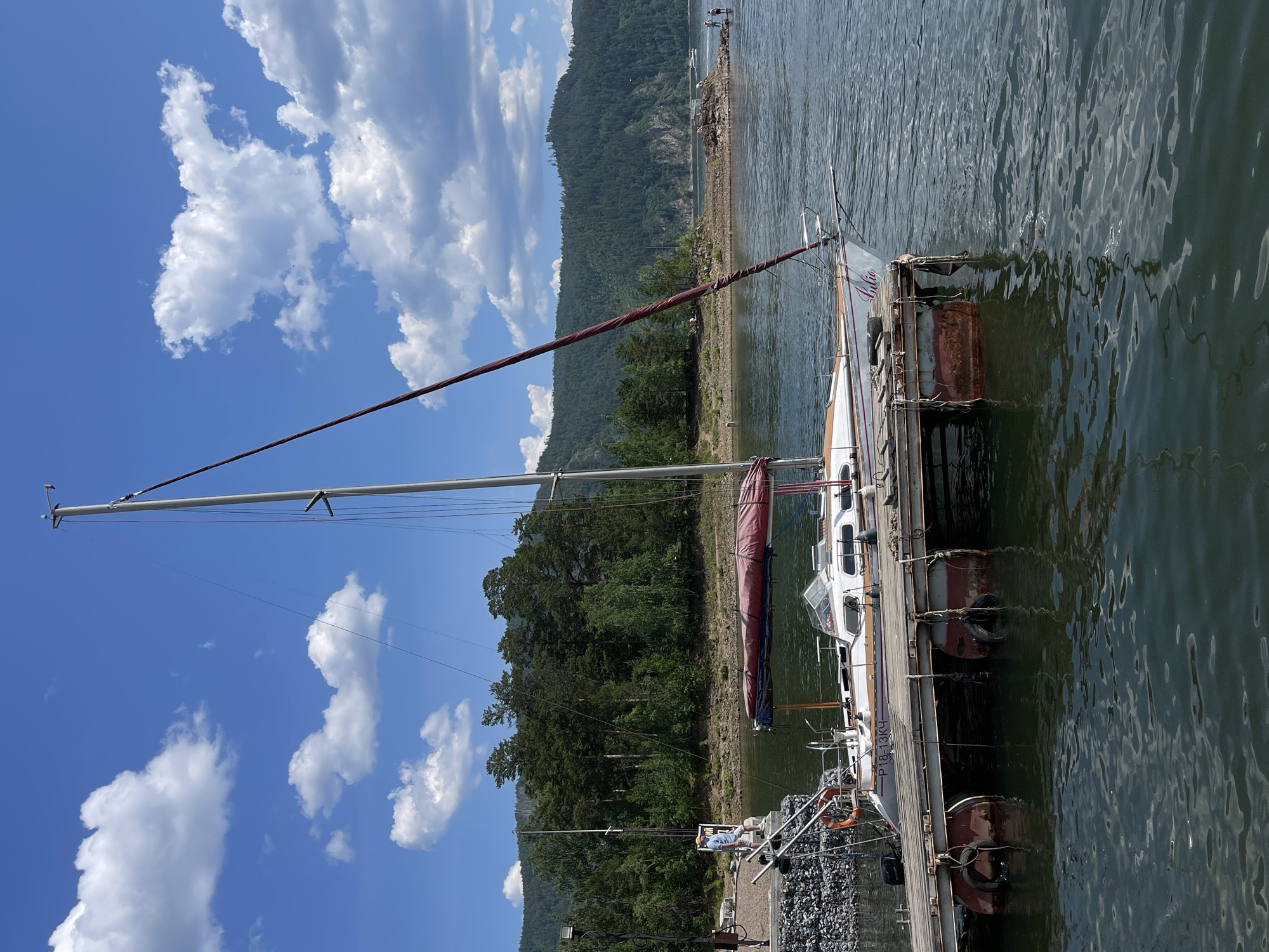 This image captures a serene scene at a dock, likely in Divnogorsk, Russia. A sailboat, with the name "Julia" visible on its side, is moored to a wooden pier. The boat is white with orange accents and has a prominent mast. The water surrounding the dock is calm and reflects the clear blue sky dotted with fluffy white clouds.
In the background, a densely forested hillside rises steeply from the shore, its green expanse meeting the clear blue sky. The time of day appears to be midday, given the bright sunlight casting shadows and the vibrant colors. The weather is fair, with a gentle breeze suggested by the rippling water.
On the dock, a person wearing a blue shirt is visible, seemingly engaged with the boat or its surroundings. A life preserver is also on the dock, a common safety feature at a marina. The dock itself is made of wood and is lined with rocky material on one side, possibly for erosion control or as a foundation.
Visible text on the boat includes "Julia" and "P18-13K4". The overall mood of the image is peaceful and tranquil, evoking a sense of leisure and connection with nature. The setting suggests a popular spot for sailing and outdoor activities on the water.
