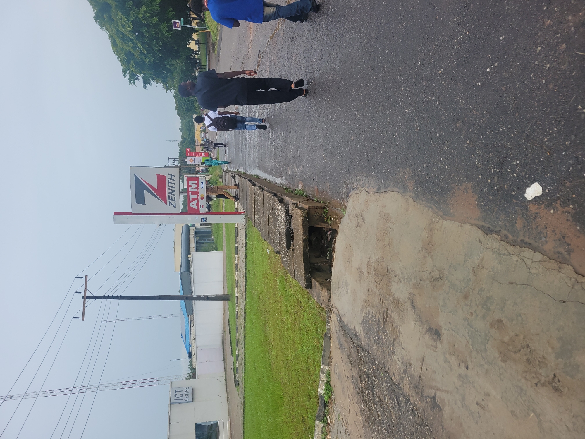 The image captures a street scene with several individuals walking on a wet asphalt road. A white building with "ICT CENTRE" signage is visible on the left. To the right of the building, a Zenith Bank ATM sign is displayed on a red and white pole. Several people are walking away from the camera along the sidewalk and the road. The environment appears to be a moderately developed urban area, with green grass, trees, and utility poles visible. The sky is overcast.
