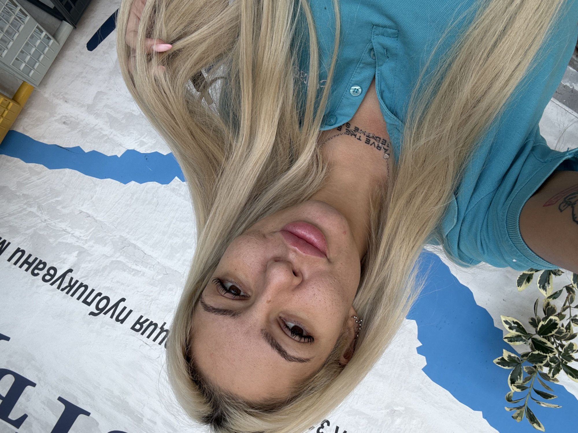 A selfie shows a blonde woman wearing a blue polo shirt, lying on a surface marked with white, blue, and illegible Cyrillic text. Some green plants are visible near her right shoulder. The location appears to be Plovdiv, Bulgaria, based on the Cyrillic text.