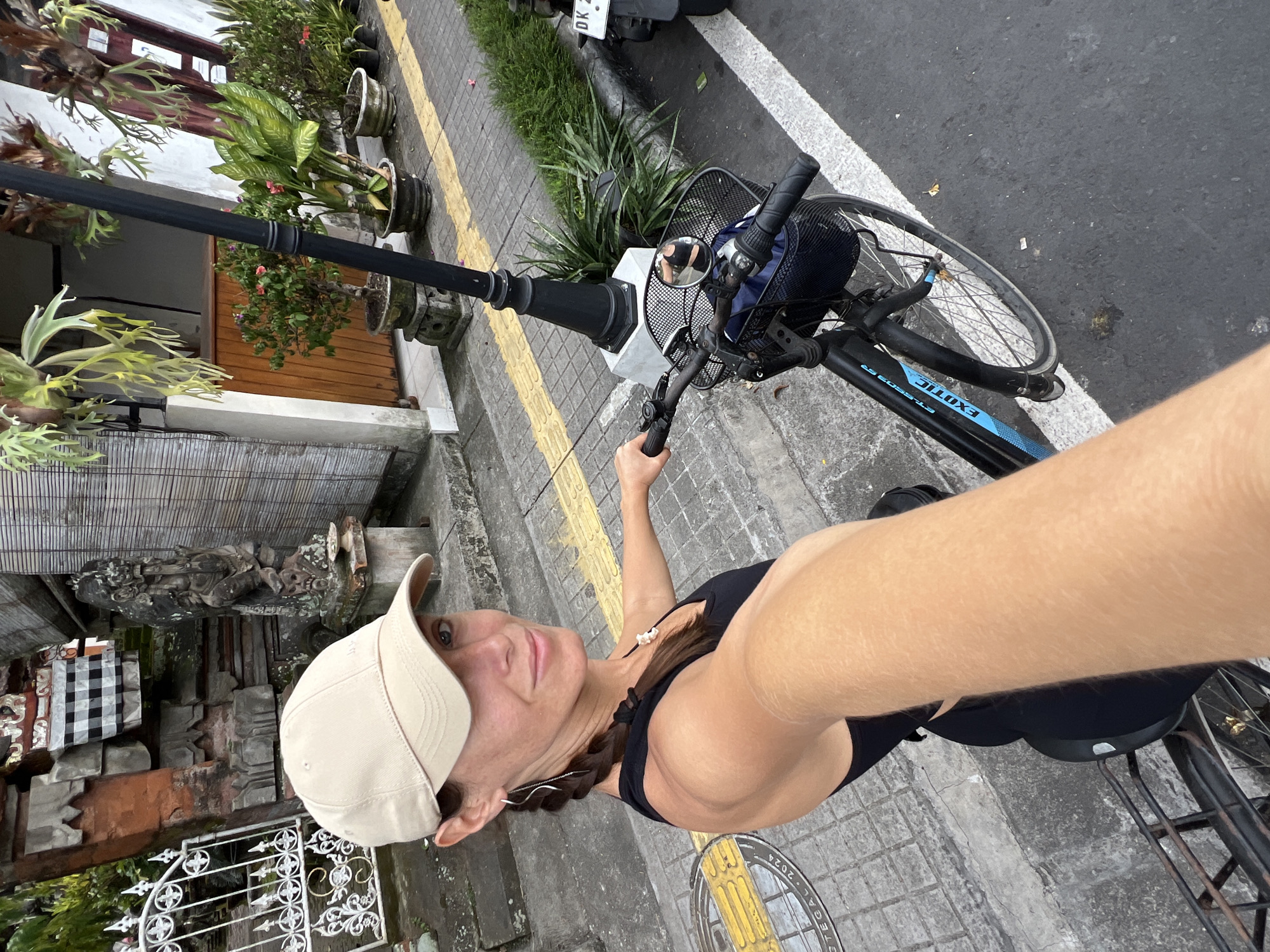 A woman wearing a baseball cap, a black tank top, and dark-colored bottoms is taking a selfie while riding a bicycle in Ubud District, Indonesia. The bicycle is black with blue accents and has a basket in the front. The woman is holding the handlebars with her right hand. The background shows a paved street with a yellow line, a lamppost, and various potted plants along a building with wooden doors and stonework. A motorcycle is partially visible parked ahead of the bicycle.