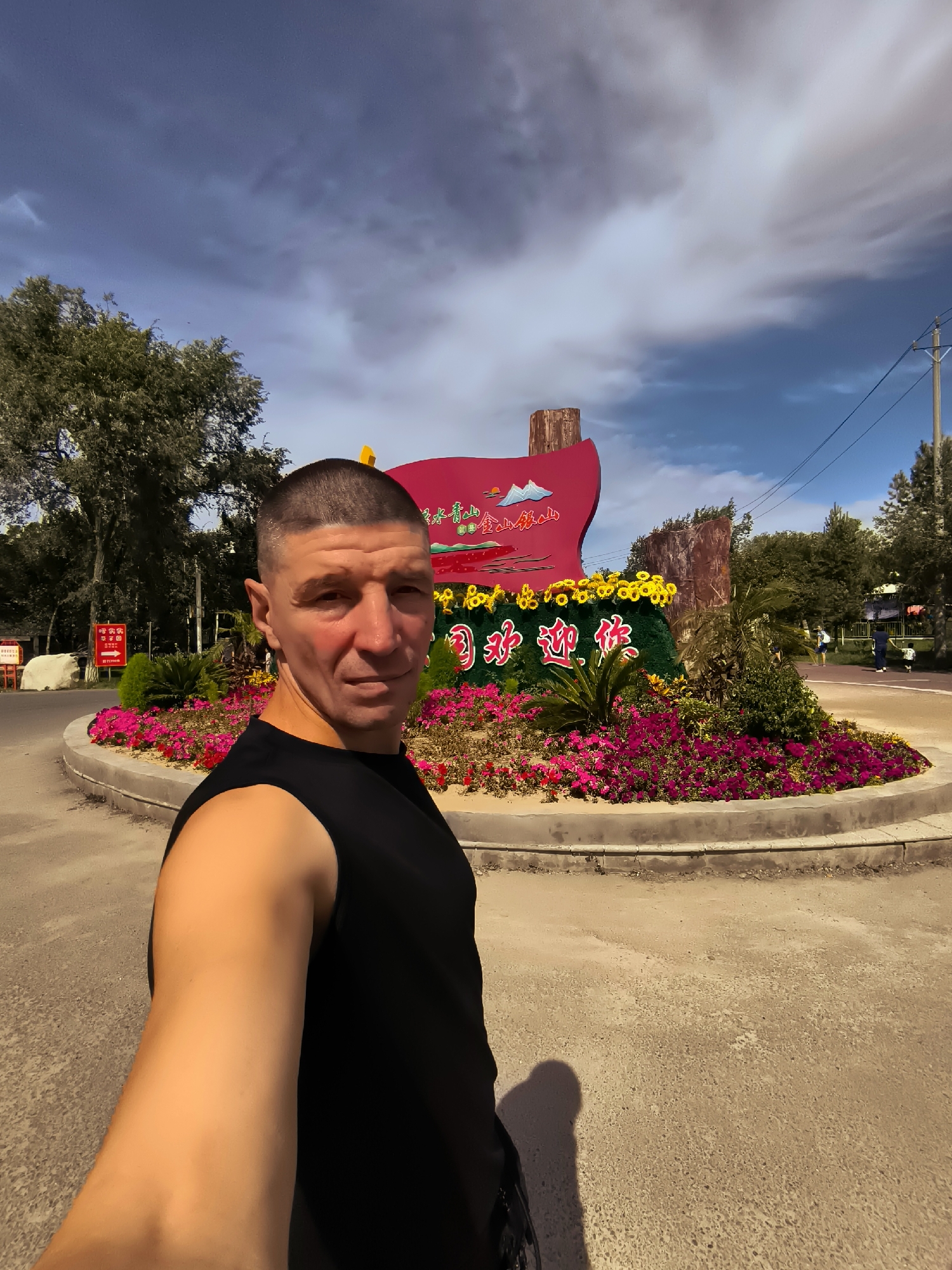 The image shows a man taking a selfie in front of a large, red sign adorned with colorful flowers and text. The sign appears to be a welcome sign, likely at an entrance to a park or scenic area. The man is wearing a black sleeveless shirt and has short, buzzed hair. He is looking directly at the camera with a neutral expression. The background features trees, a cloudy sky, and a paved area that could be a road or walkway. There are also hints of other people in the distance, suggesting a public space. The overall scene is set outdoors, possibly in a city park or a tourist attraction in Ürümqi, China, given the presence of Chinese characters on the sign.