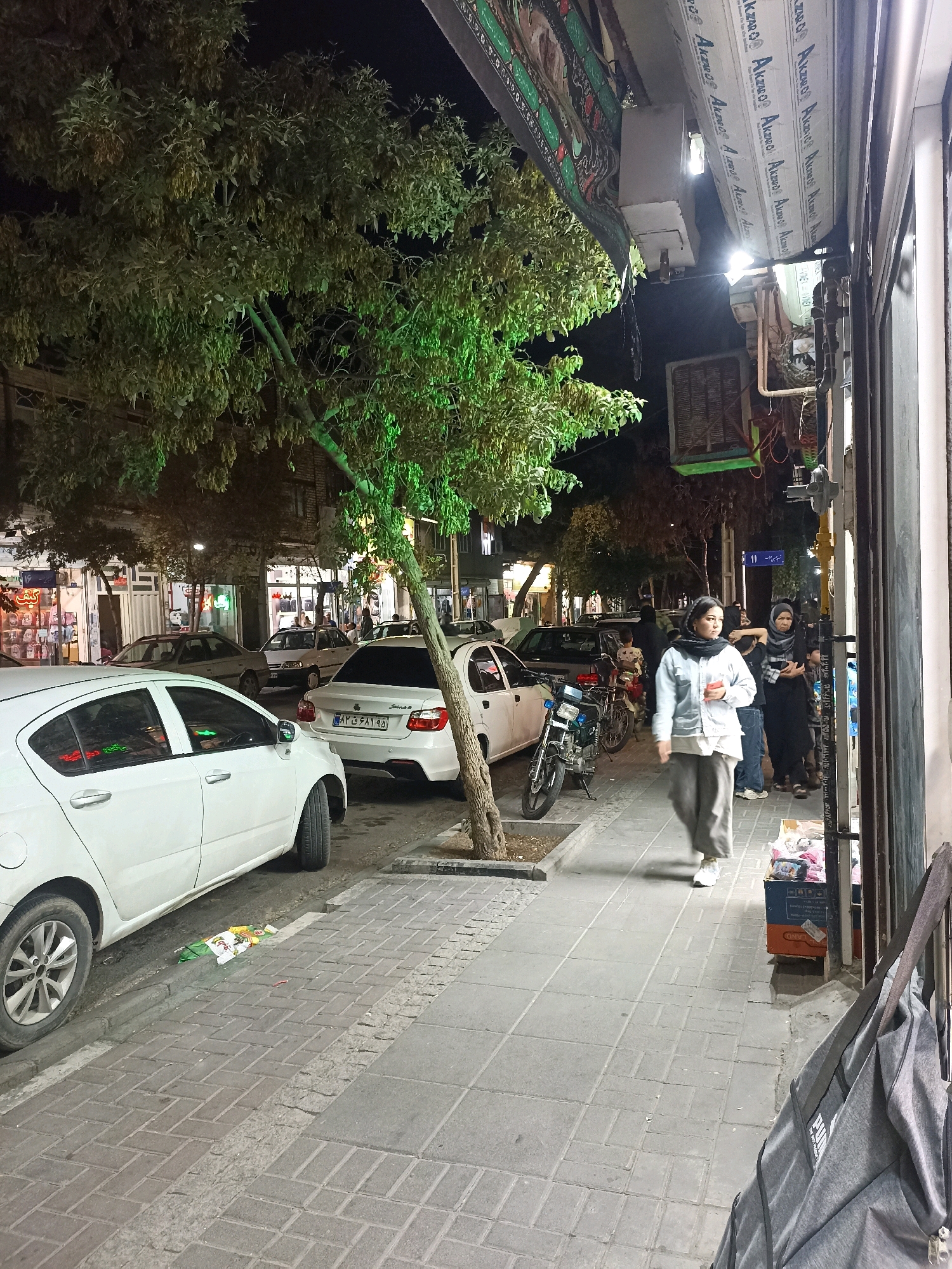 The image shows a street scene at night in Mashhad, Iran. Several white cars are parked on the side of the street. A sidewalk is visible, with a few people walking on it. One woman, wearing a light blue jacket, grey pants, and a black scarf, is walking towards the camera. Another woman, wearing a black headscarf, is standing next to her, facing away from the camera. There are shops on the right side of the street. A tree with green leaves is in the foreground. The street is paved with bricks.