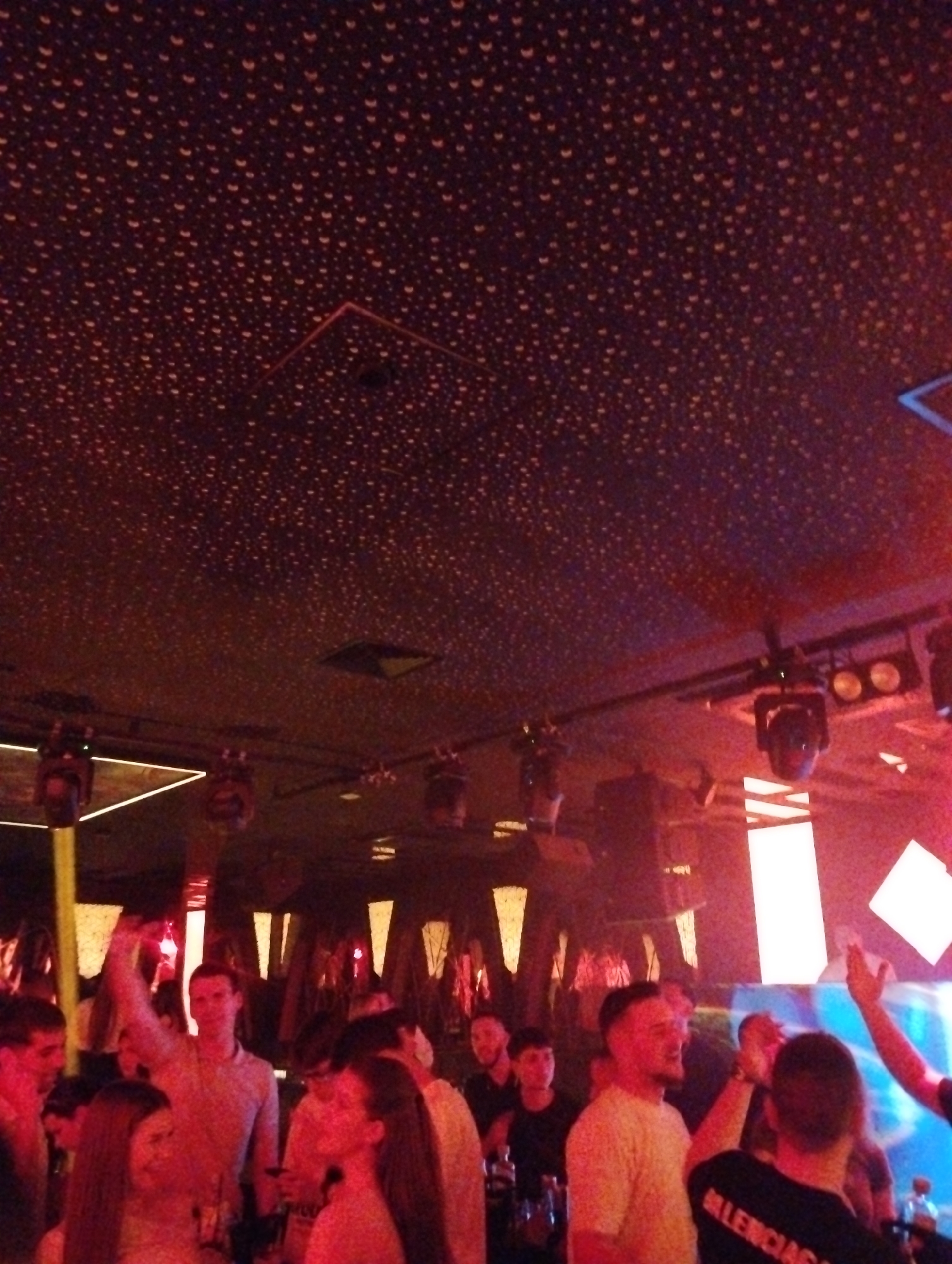 Here is a description of the image:
The image shows a crowded nightclub scene. Many people are standing close together. Several individuals are visible, including men wearing t-shirts and a woman with long hair. Overhead, there are several stage lights and decorative panels, along with a dark ceiling covered with small, illuminated dots. The lighting creates a strong red tint to the scene. The word "Balenciaga" is visible on the back of a black t-shirt. The location and city are unknown.