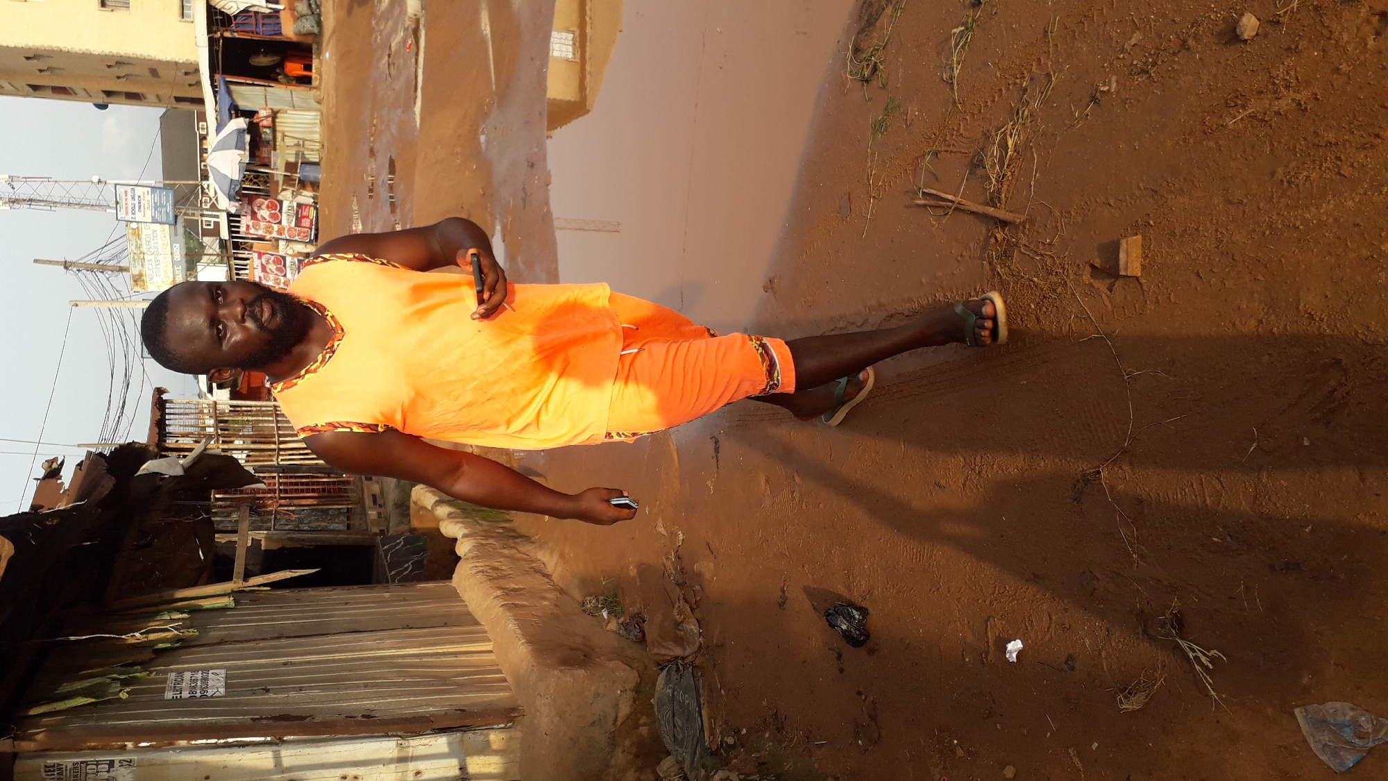 A man stands in a muddy street in Nkpor, Nigeria. He wears an orange tank top and matching shorts, with sandals on his feet. He holds a mobile phone in each hand. The environment includes puddles, brown dirt, trash, and a dilapidated corrugated metal structure to the left. Buildings are visible in the background. Overhead wires crisscross the sky.